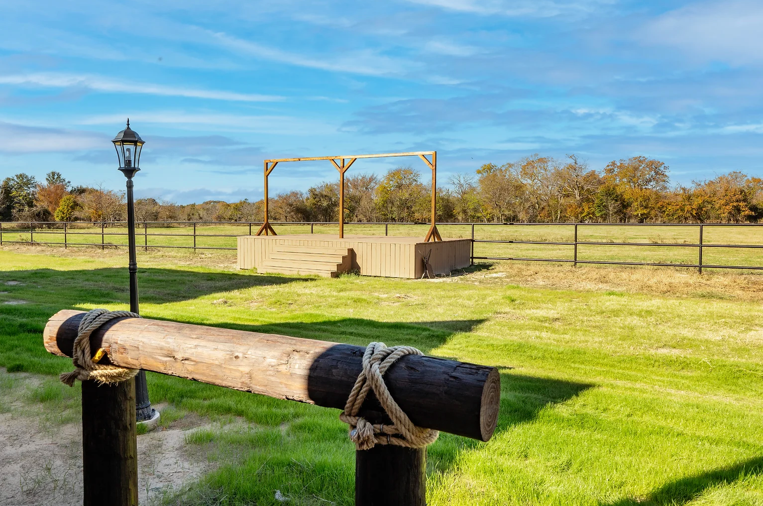 A wooden gallows structure stands in a grassy field under a sunny sky, viewed past a decorative log railing.