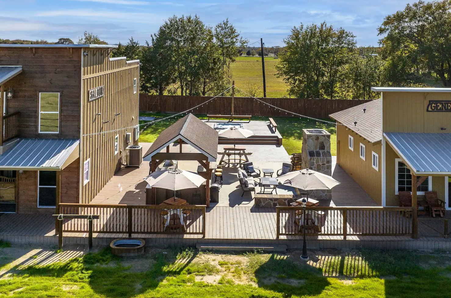 An elevated view of a rustic wooden patio between two buildings, featuring outdoor seating, umbrellas, and string lights.