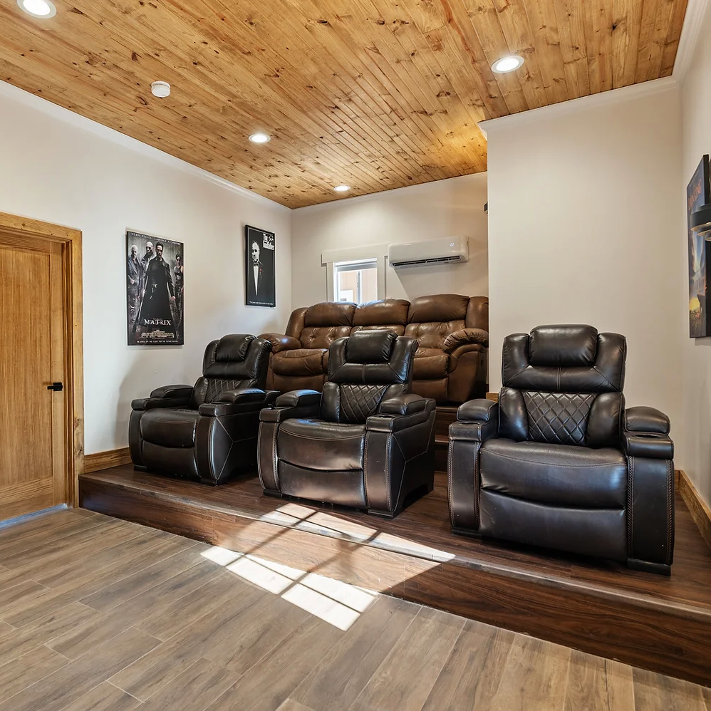 A home theater room with three dark leather recliner chairs on a raised platform, wood-planked ceiling, and wall posters.
