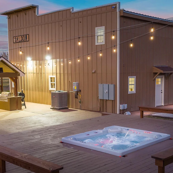 A hot tub on a wooden deck outside a barn-style building with string lights at dusk.