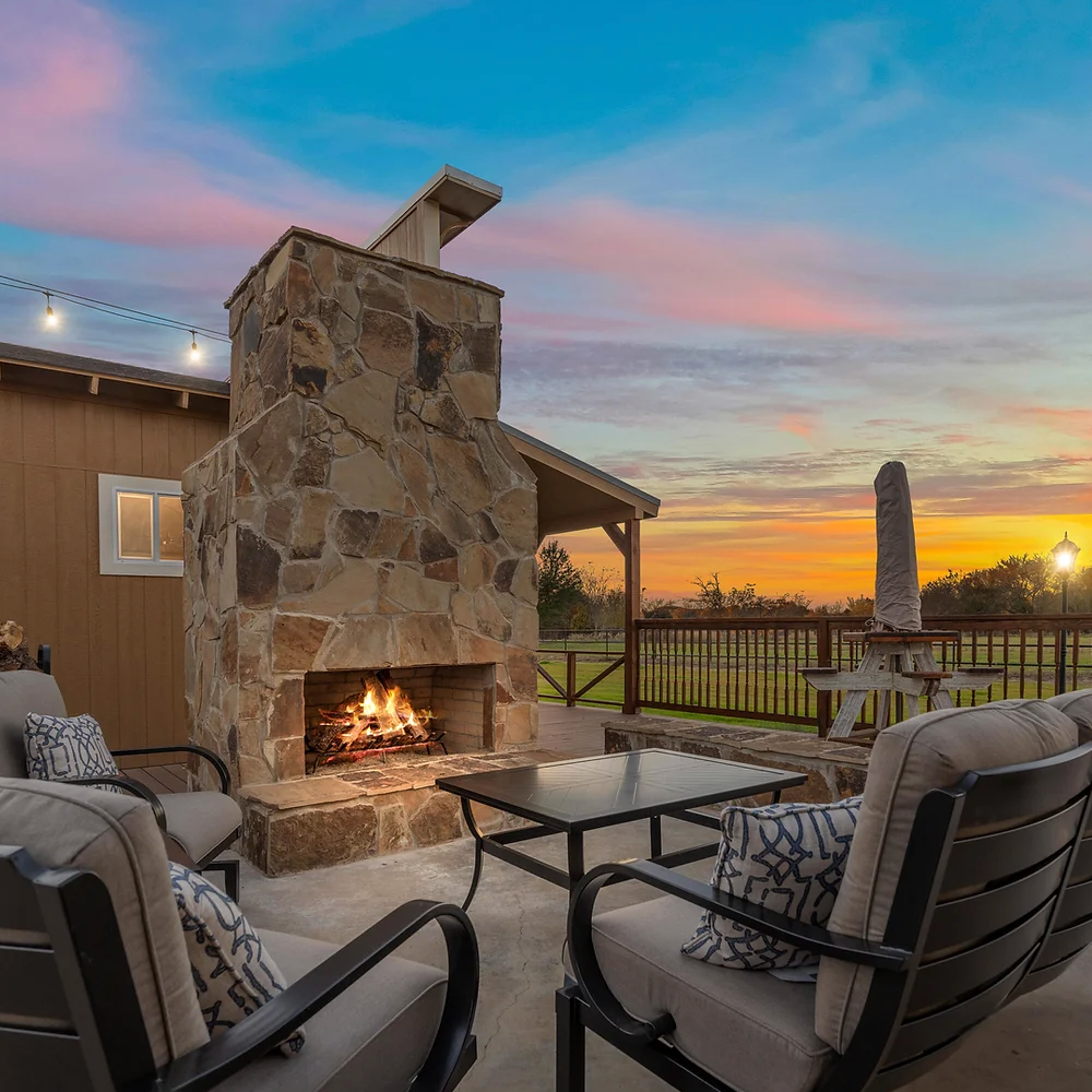 An outdoor stone fireplace on a patio at sunset with cushioned chairs, a coffee table, and string lights.