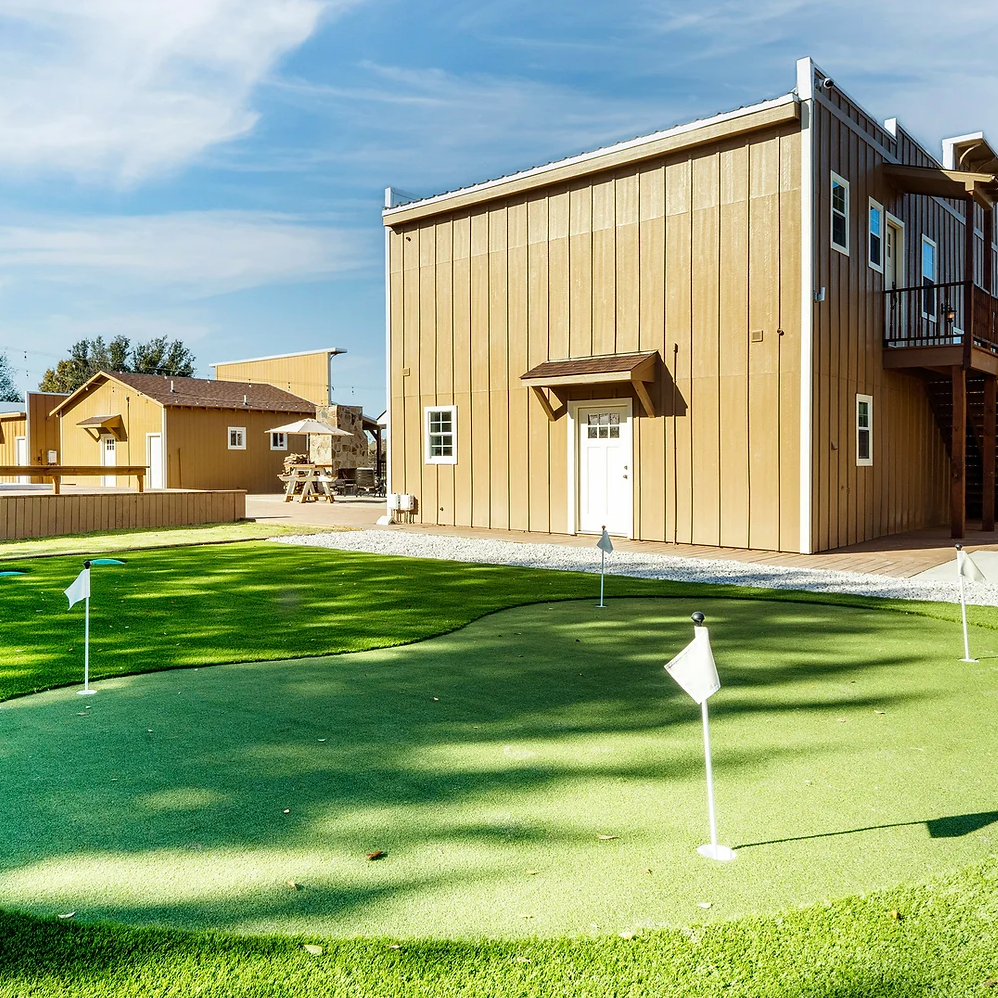 A green putting area with golf flag pins sits in front of tan buildings under a clear blue sky.