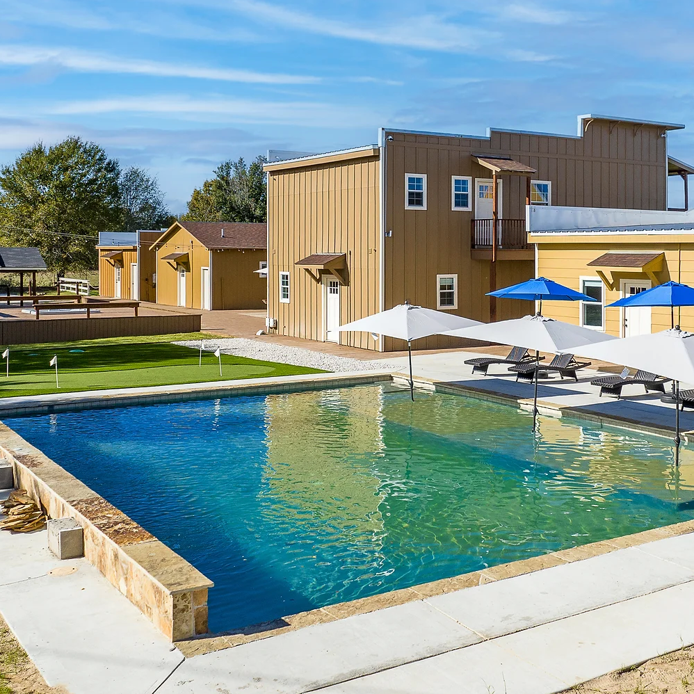 A rectangular turquoise swimming pool sits in front of tan buildings with white umbrellas on a sunny day.