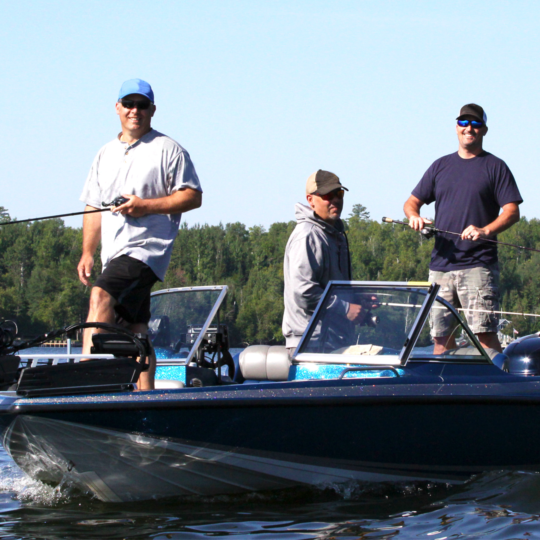 Three people fishing from a blue boat on a lake in front of a forested shoreline.