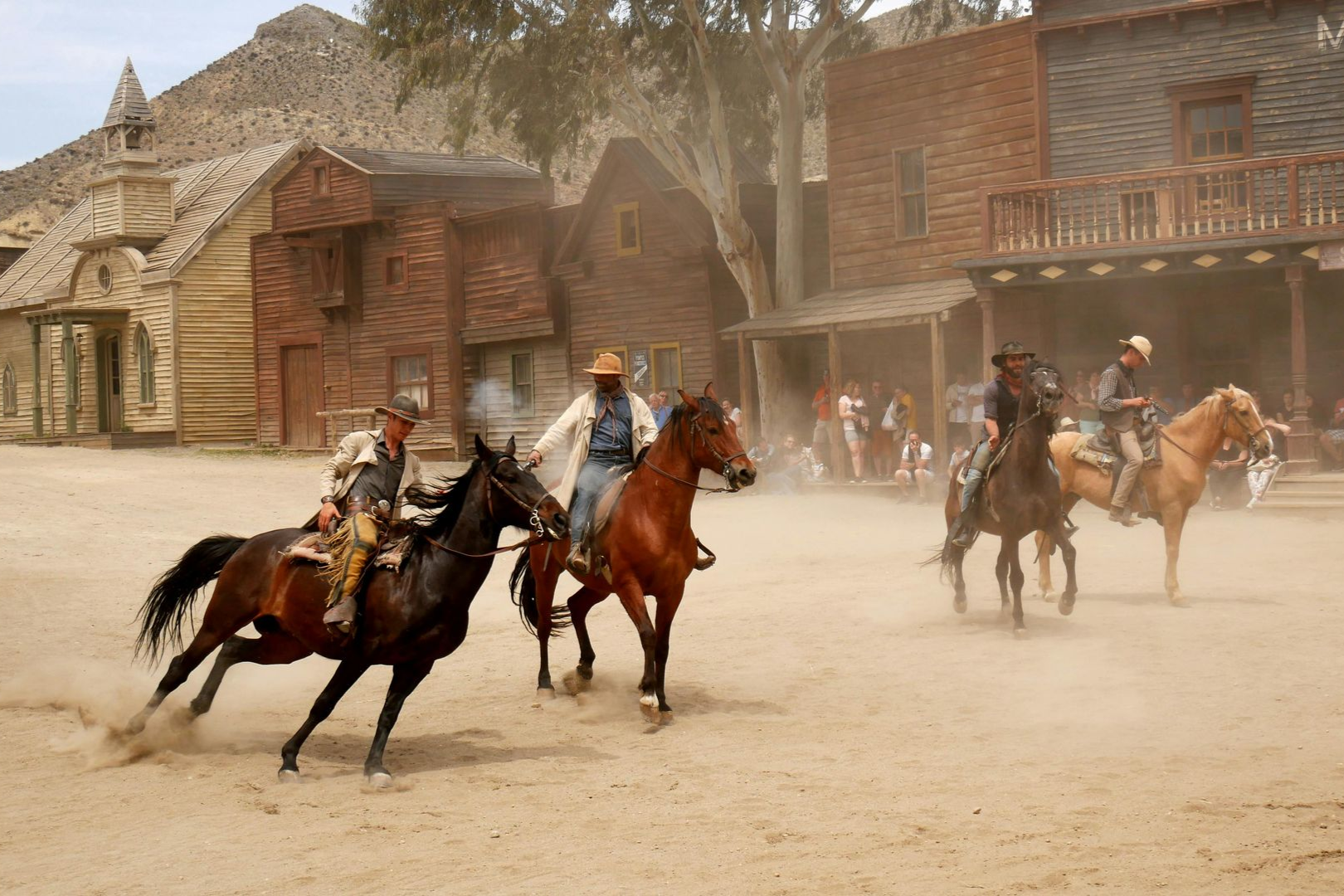 Four people on horseback ride across a dusty town square with wooden buildings in the background.