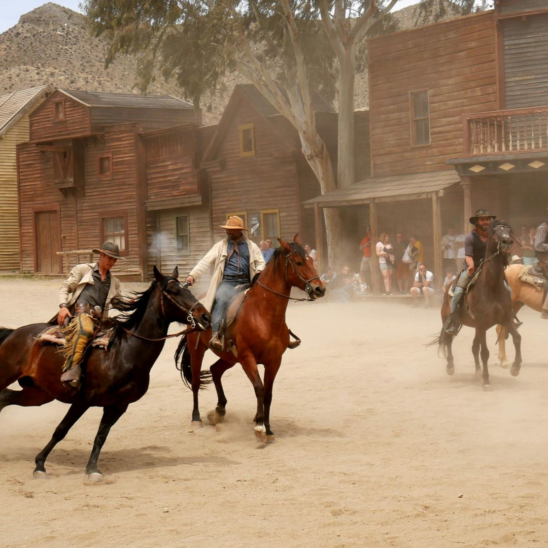 Cowboys on horseback racing through a dusty Old West street with wooden storefronts in the background