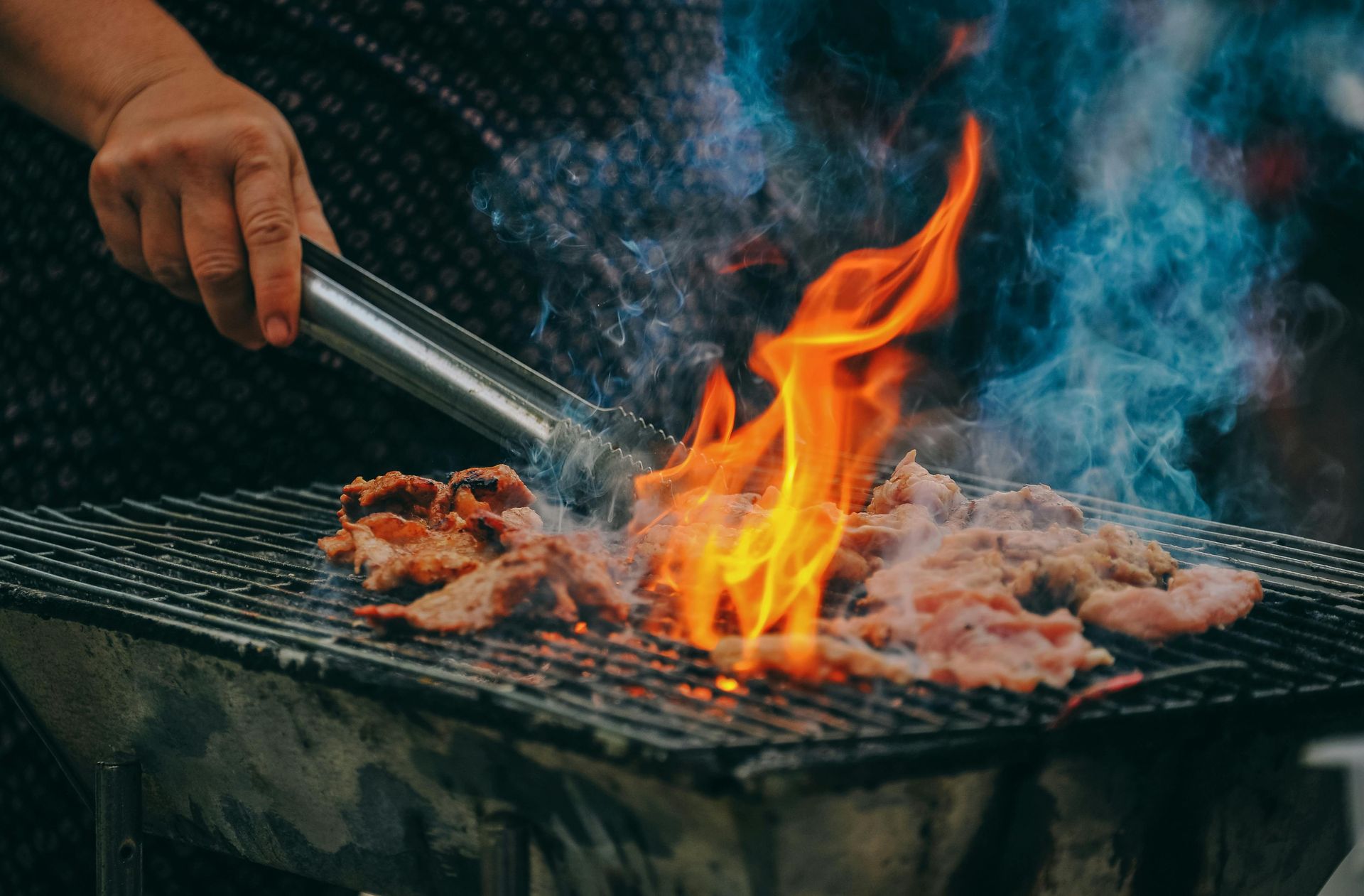 A hand uses metal tongs to grill small pieces of meat over an open flame on a charcoal barbecue.