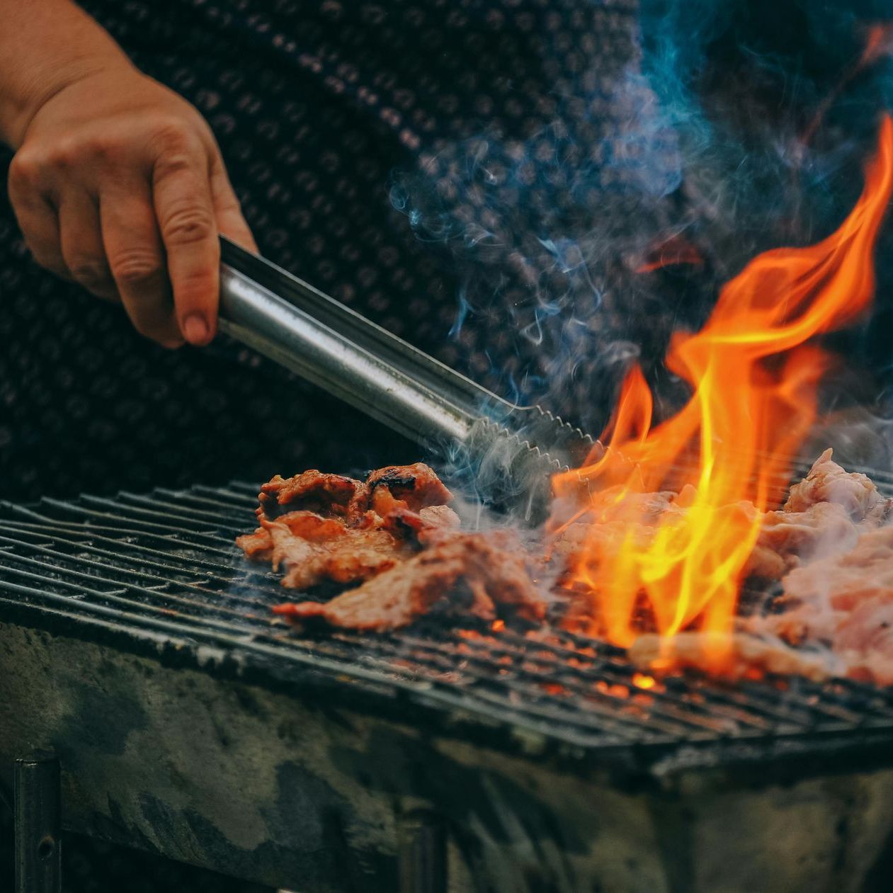 Hand using metal tongs to grill meat over open flames on a barbecue grate