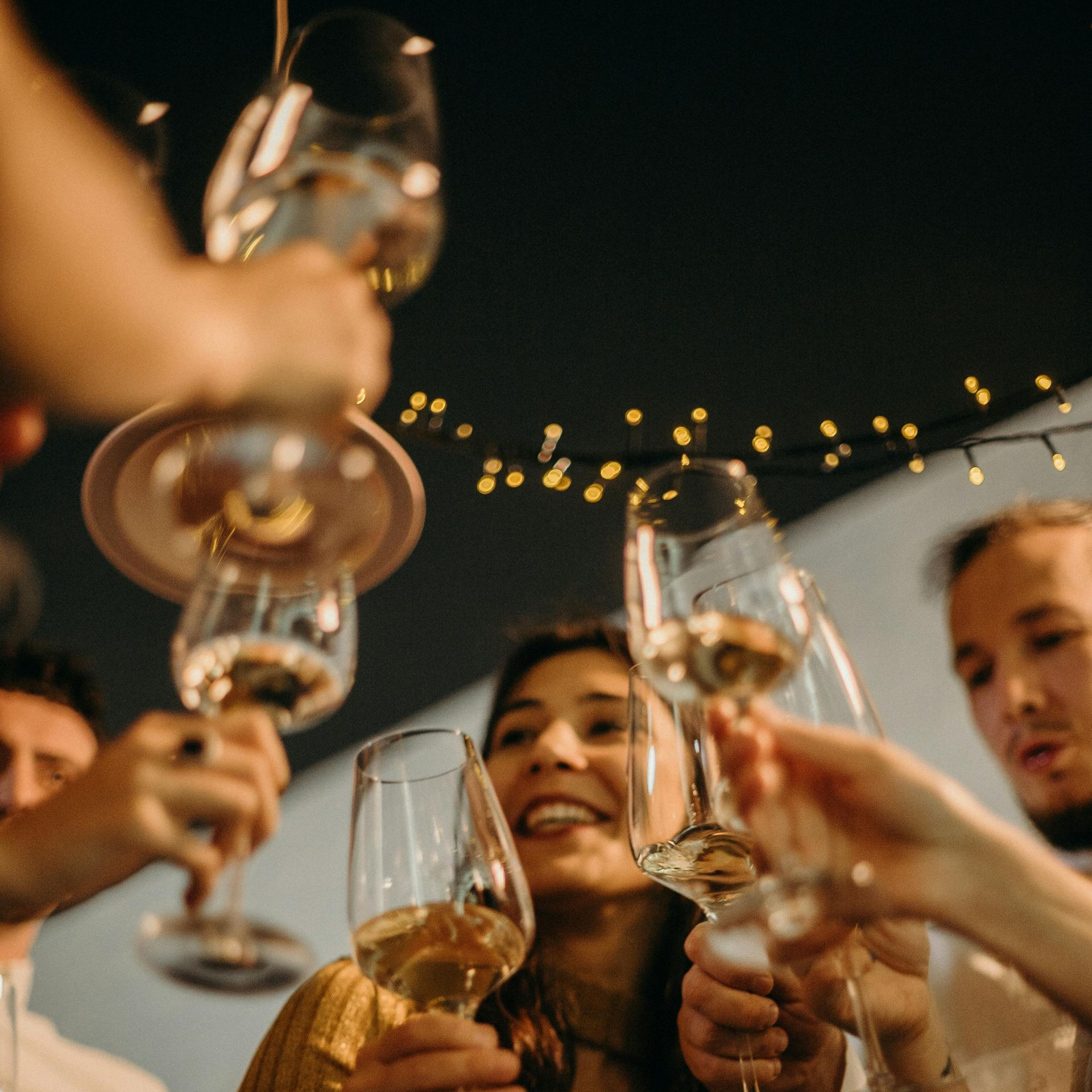 People holding wine glasses for a toast against a backdrop of festive string lights at night.