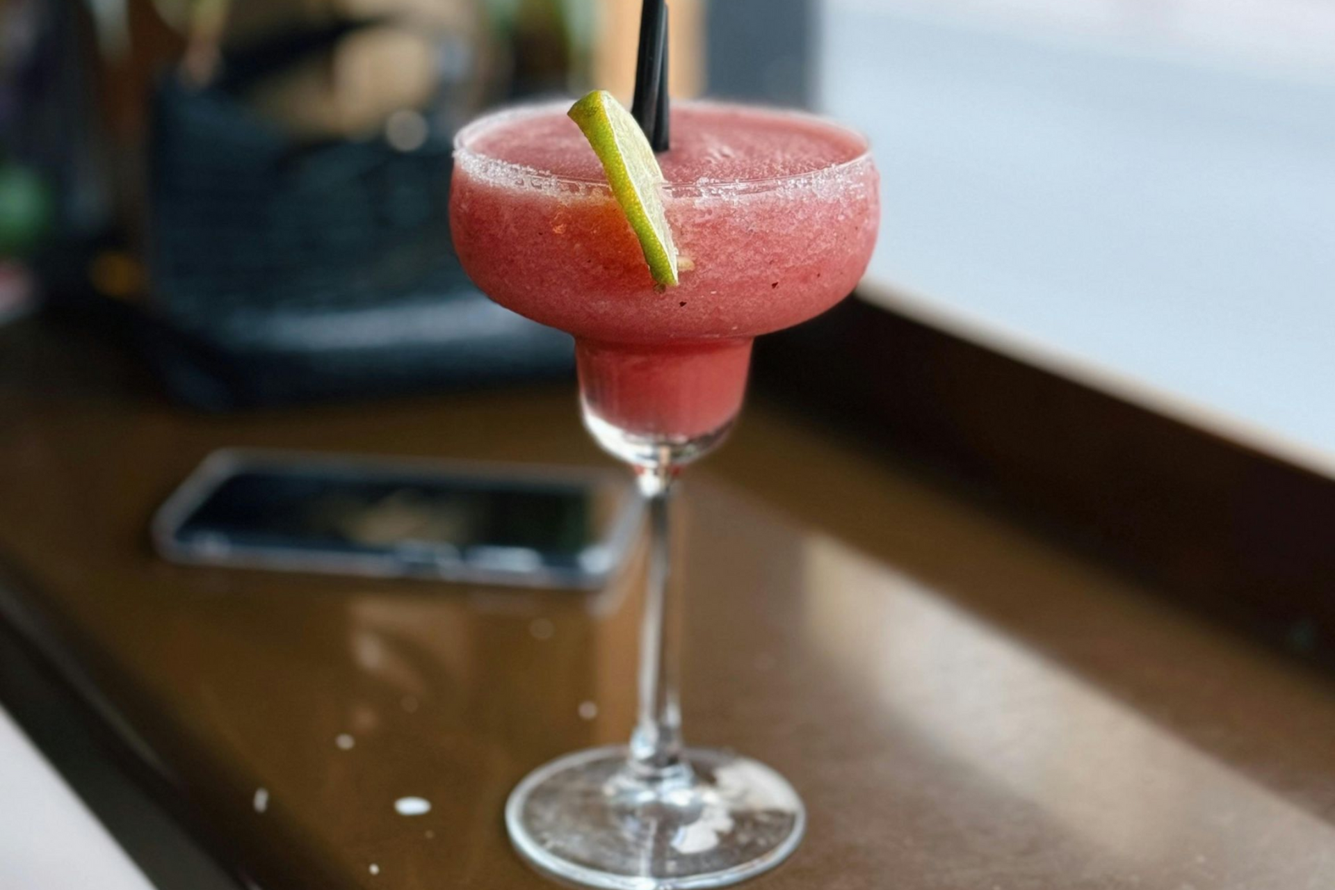A vibrant pink frozen cocktail in a rimmed glass with a lime wedge garnish, sitting on a dark counter by a window.