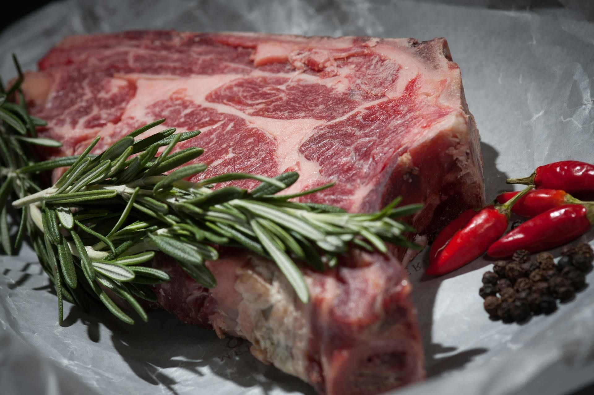 A raw T-bone steak on butcher paper with fresh rosemary, small red chili peppers, and black peppercorns.