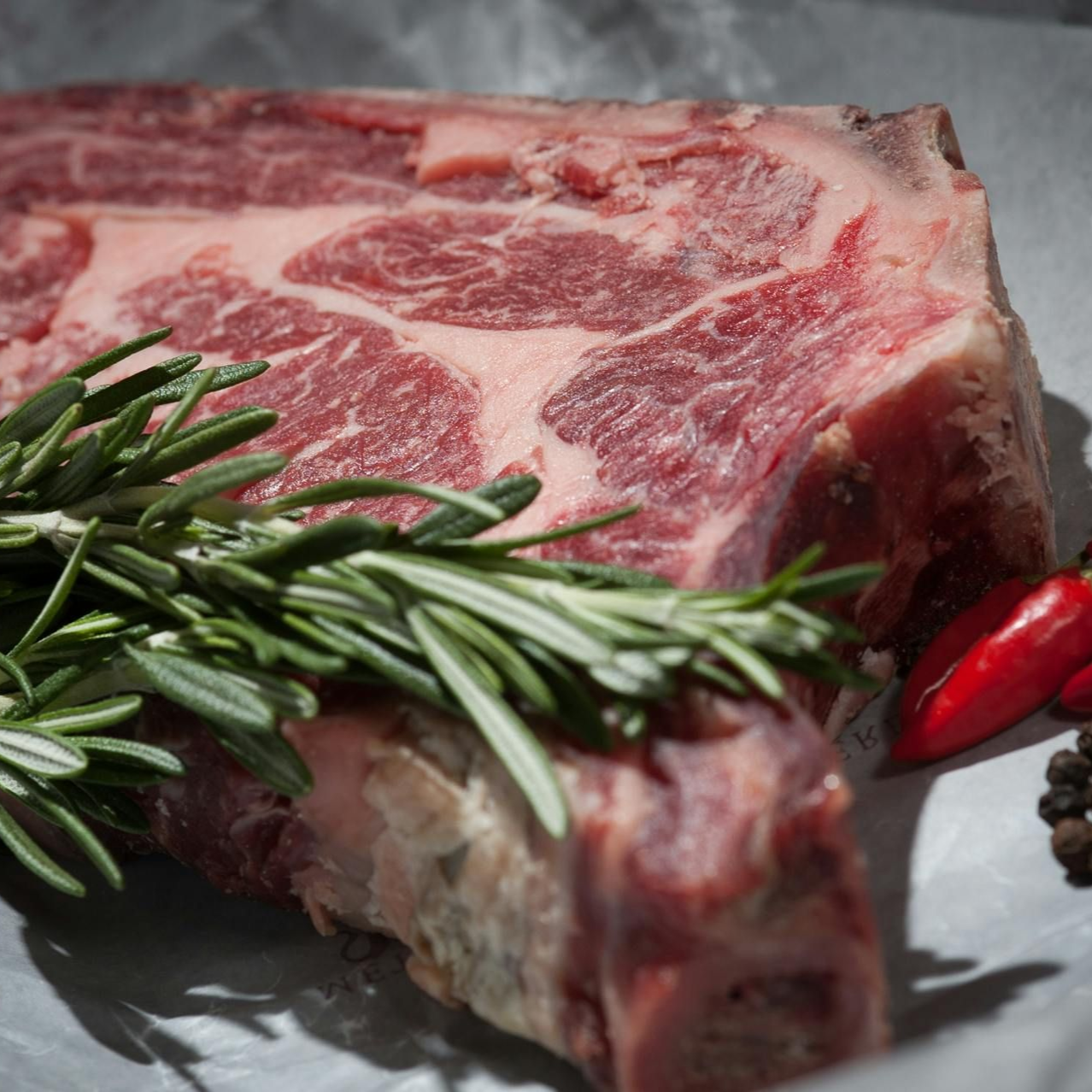 Raw marbled steak topped with rosemary, with red chili peppers on a gray surface