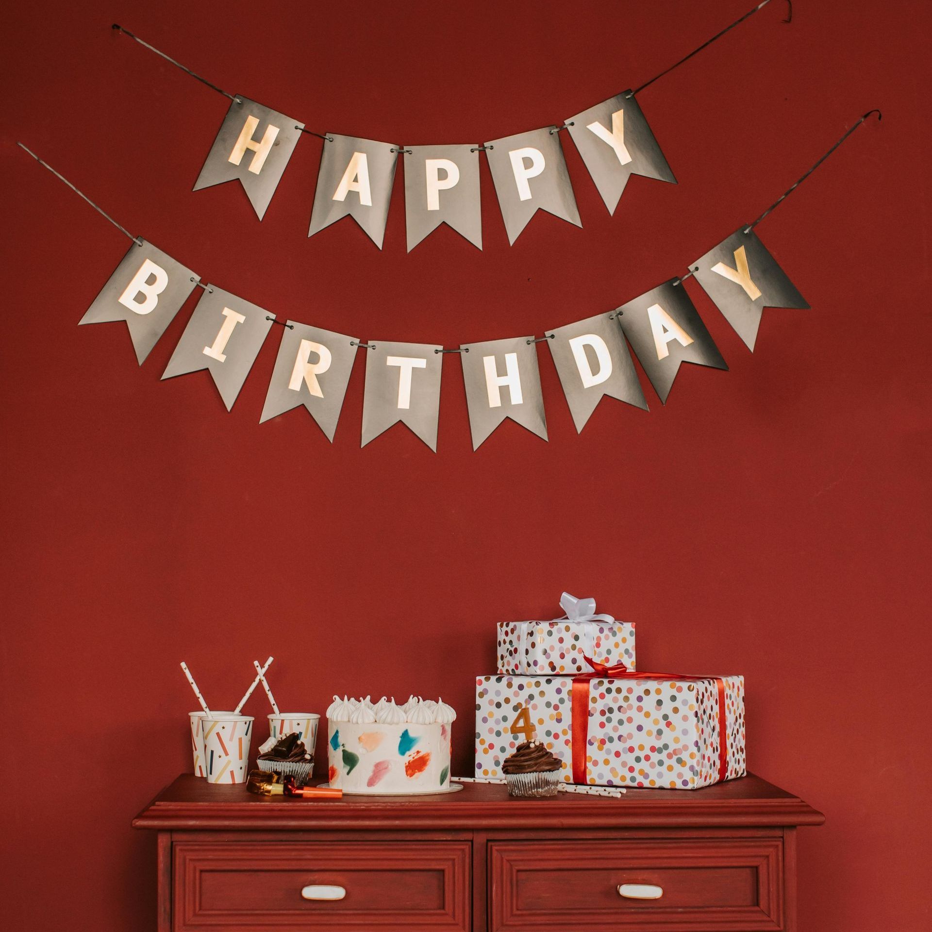 Happy birthday banner over a red wall with wrapped gifts on a wooden dresser.