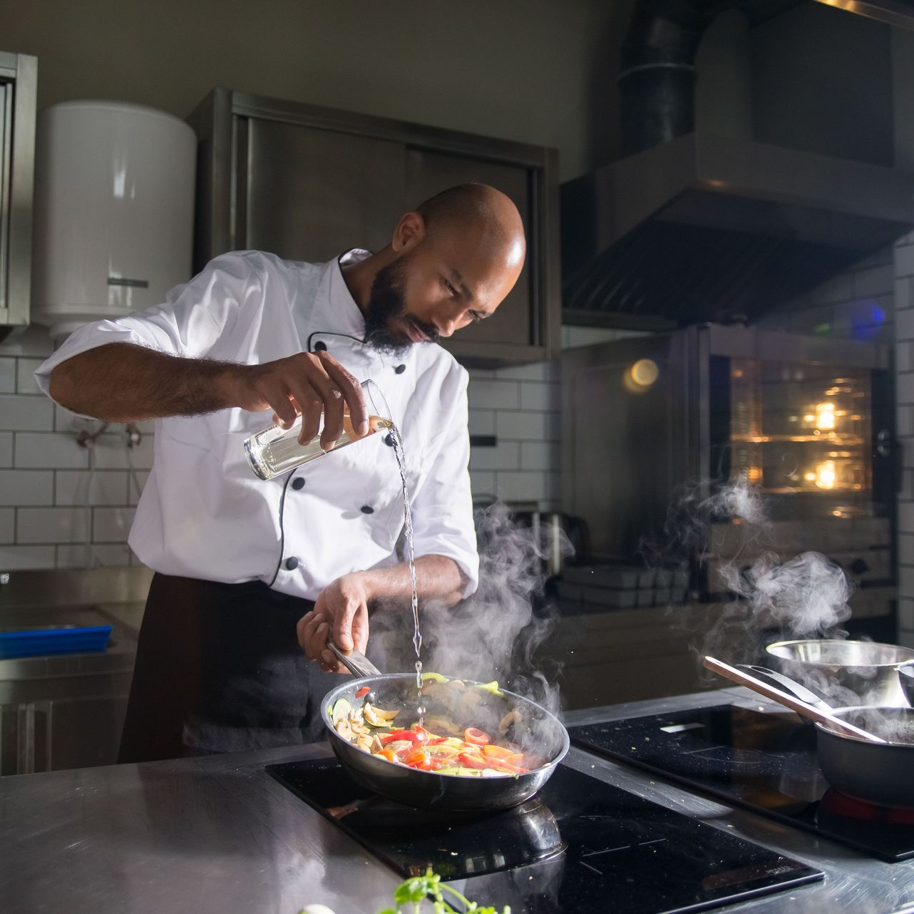 Chef seasoning a steaming pan in a modern kitchen, with another pan cooking nearby.