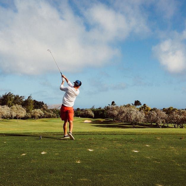 A golfer in a white long-sleeve shirt and orange shorts follows through on a swing on a sunny, grassy golf course.