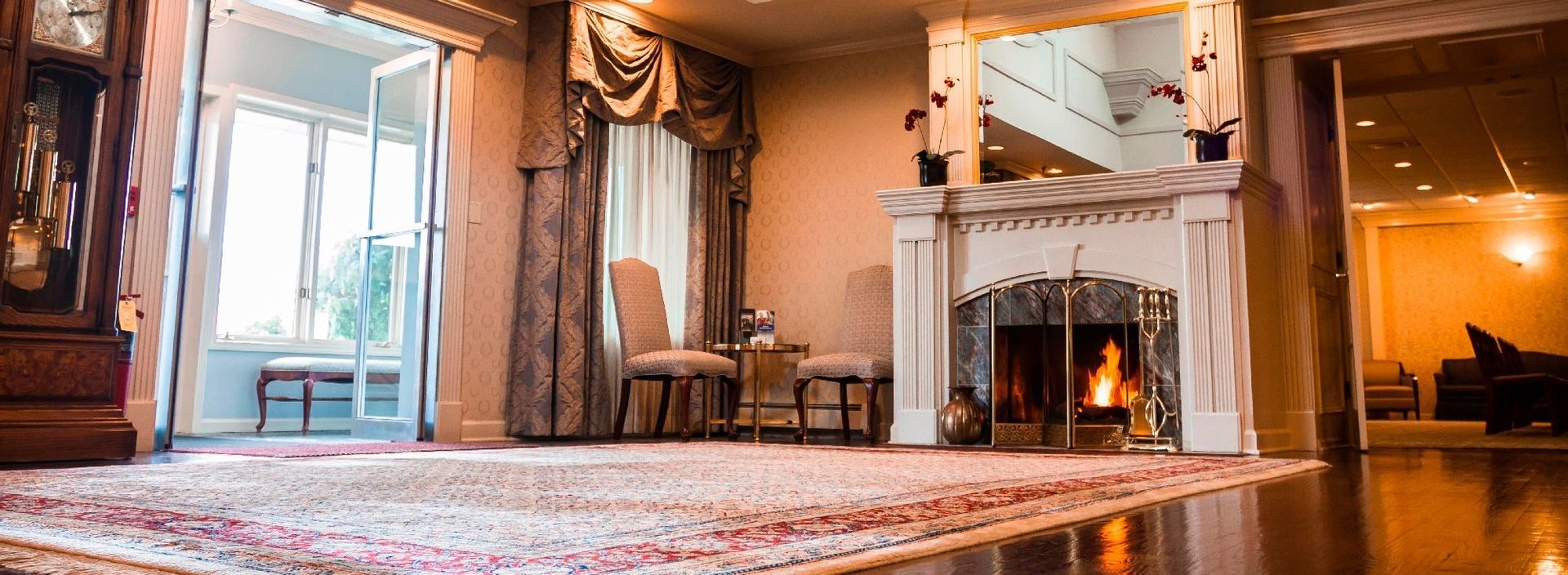 Cozy living room with a lit fireplace, large windows, and patterned rug in warm tones