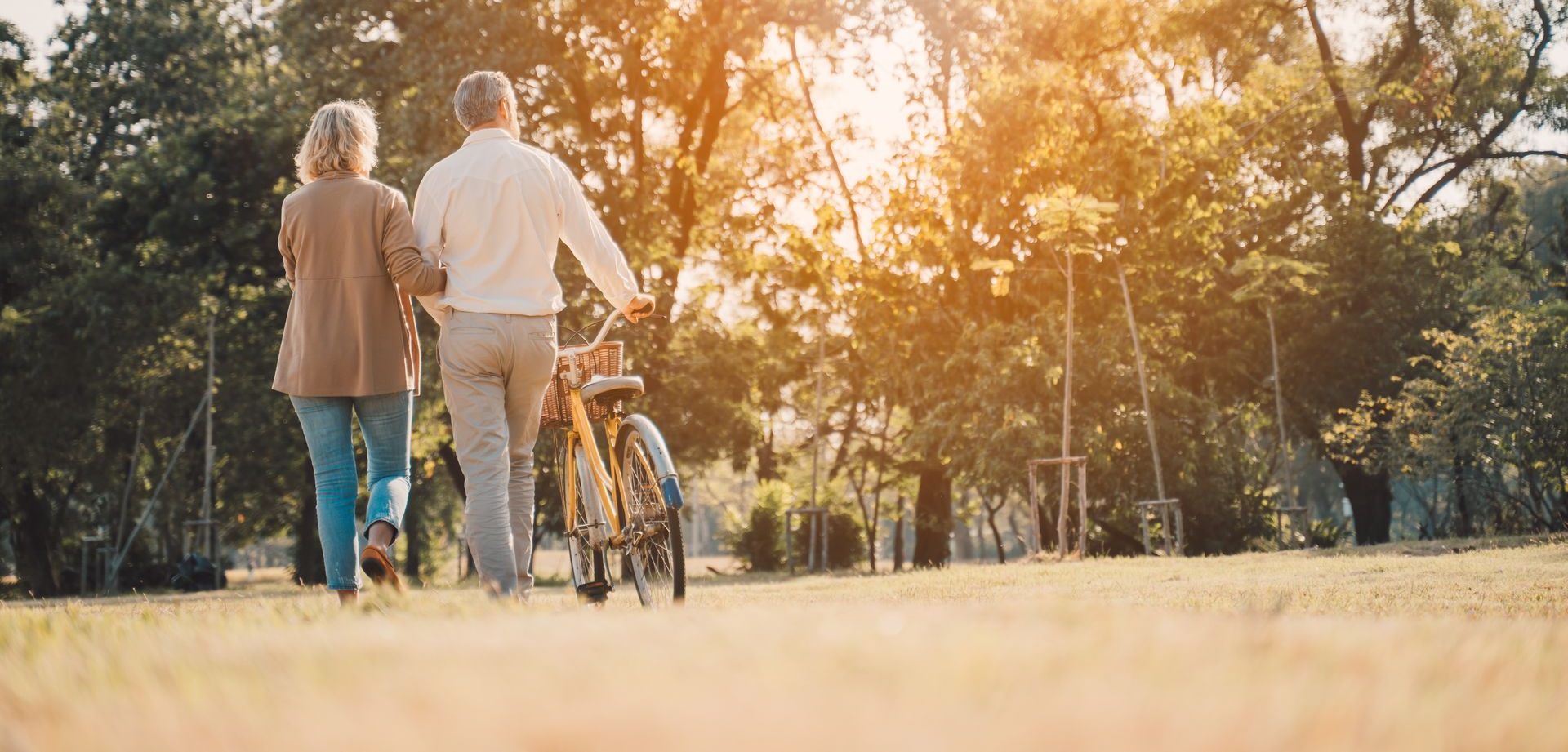 Two people walking a bicycle through a sunlit park at sunset