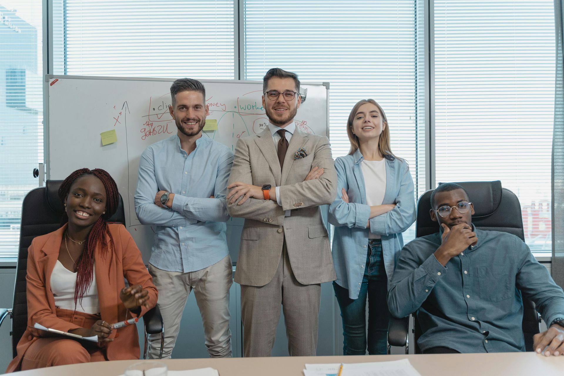 Office team poses: man in suit center, others smiling, arms crossed, in front of a whiteboard.