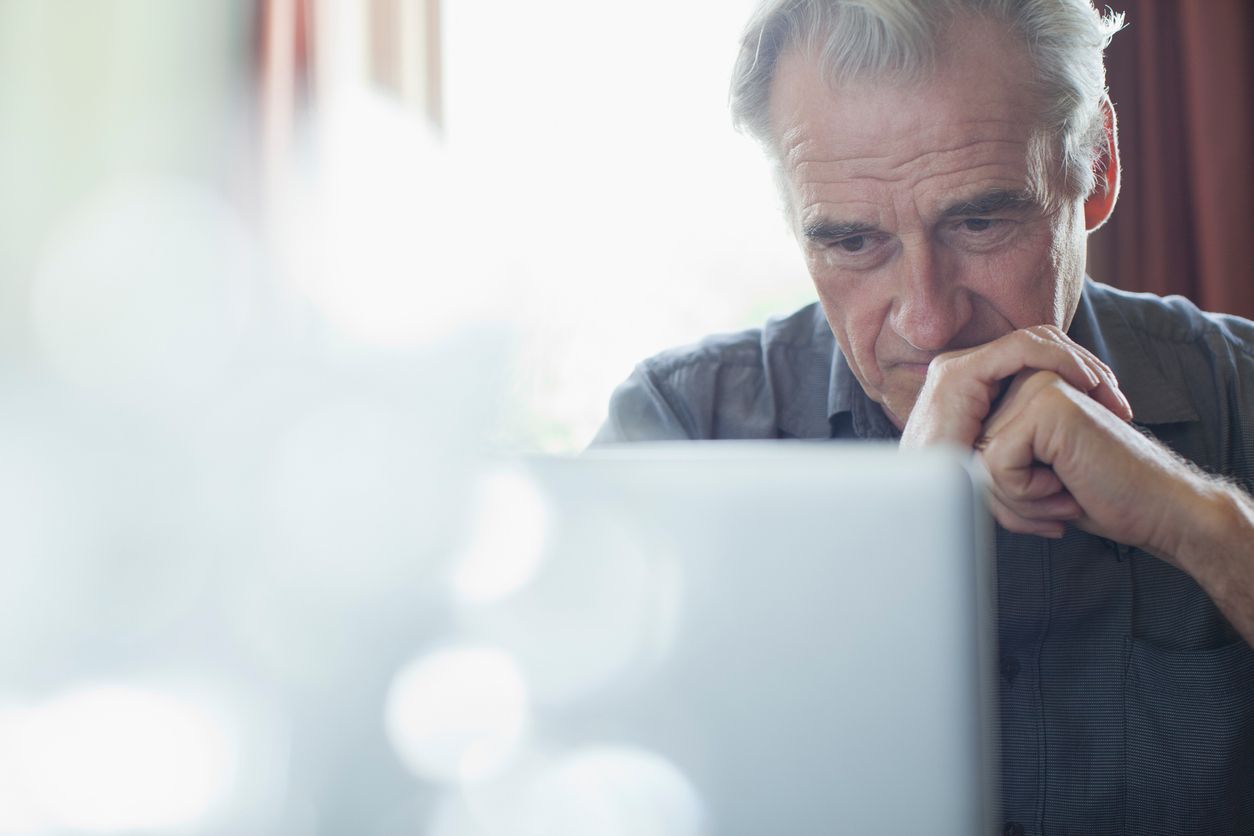 Thoughtful man looking at a laptop, resting his chin on his hand in a bright room