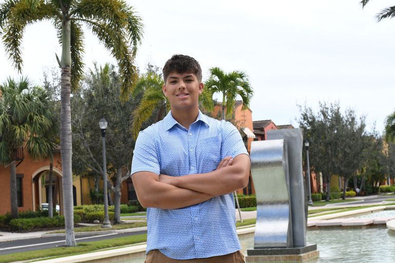 Person standing with arms crossed near a fountain in a palm-lined courtyard.