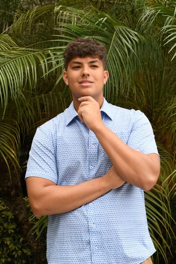 Man in a blue patterned shirt standing before lush green palms, hand on chin, looking at the camera