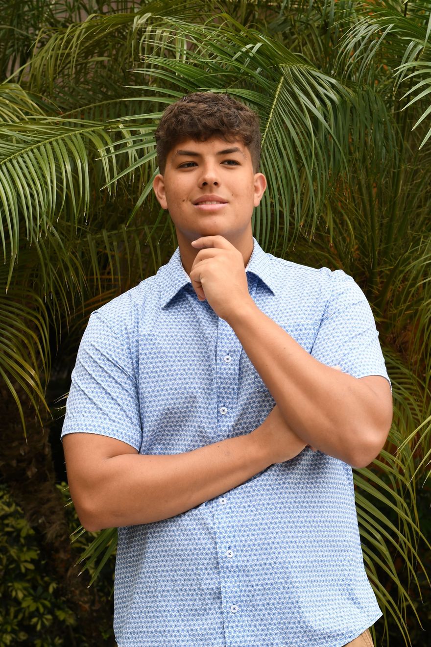 Man in a blue patterned shirt standing before lush green palms, looking at the camera