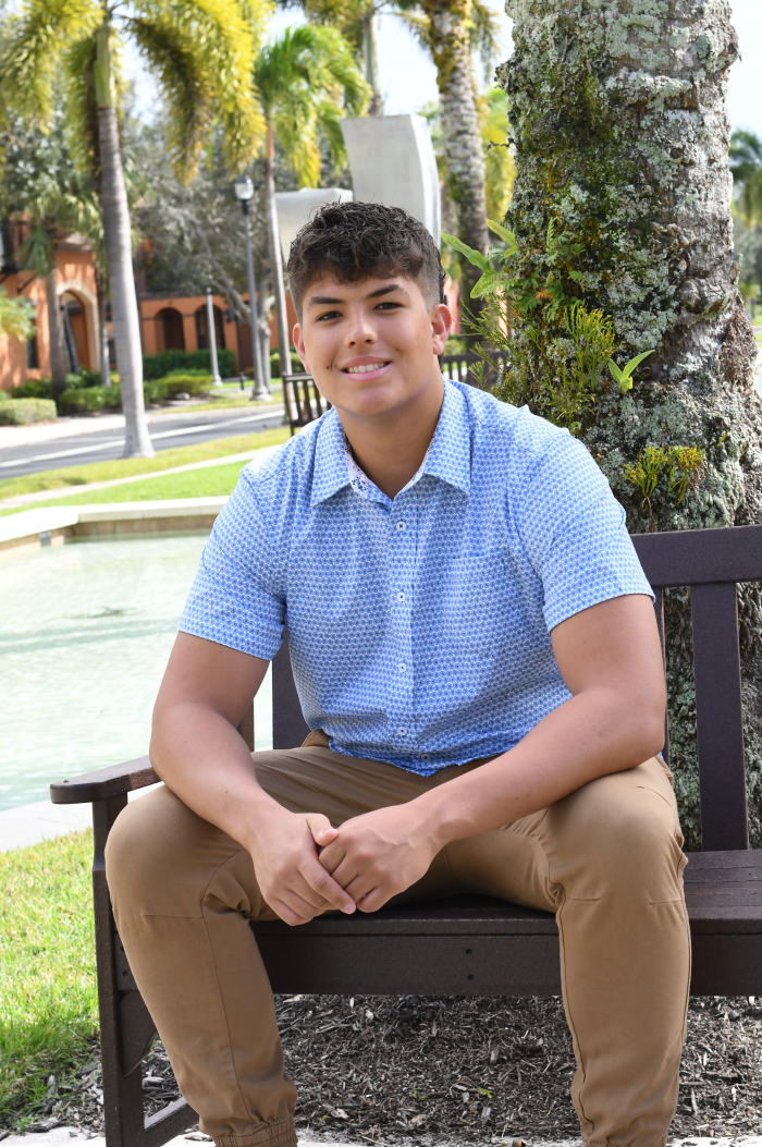 Young person sitting on a bench outdoors in a blue patterned shirt and tan pants, smiling softly.