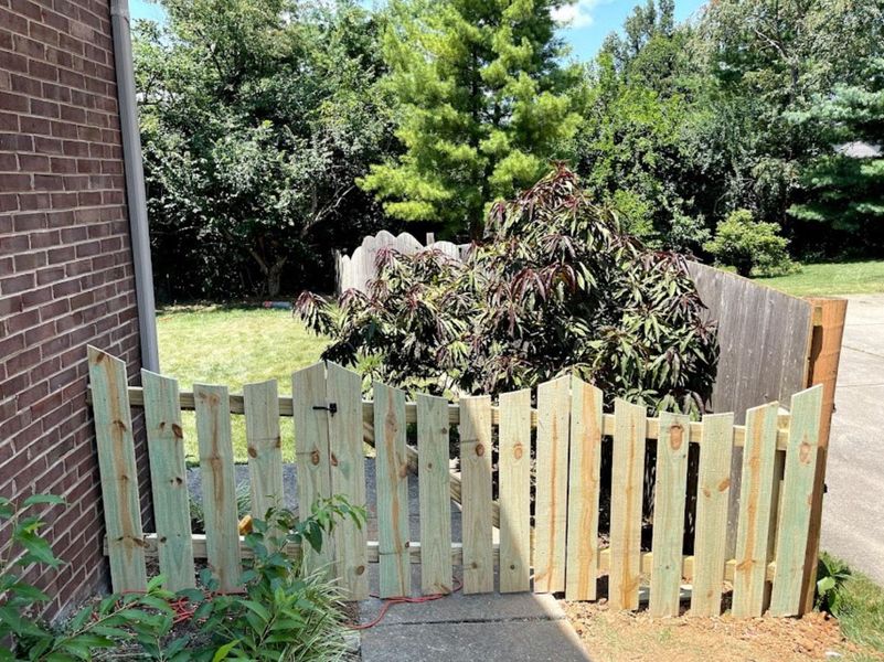 Wooden picket fence next to a brick wall. Green-tinted wood. Trees and grass in the background.
