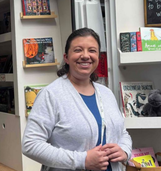 Smiling person standing in a bookstore aisle beside shelves of books and displays
