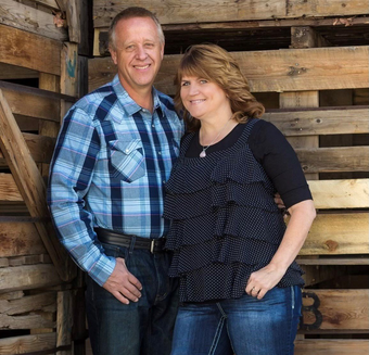 Two smiling people standing together in front of a rustic wooden wall, wearing casual blue jeans and plaid.