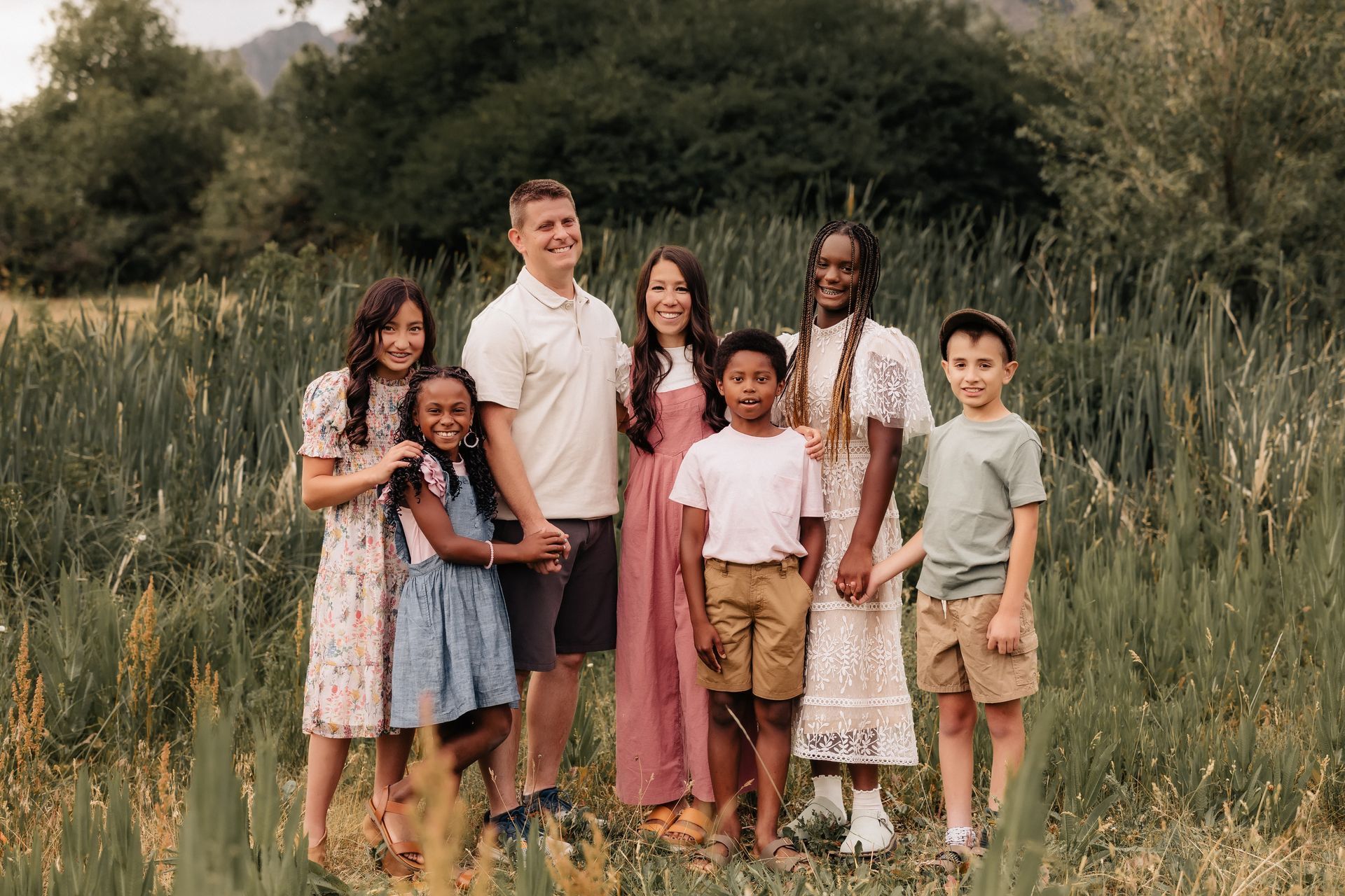 Group of seven standing in a grassy field, smiling in casual summer clothes.