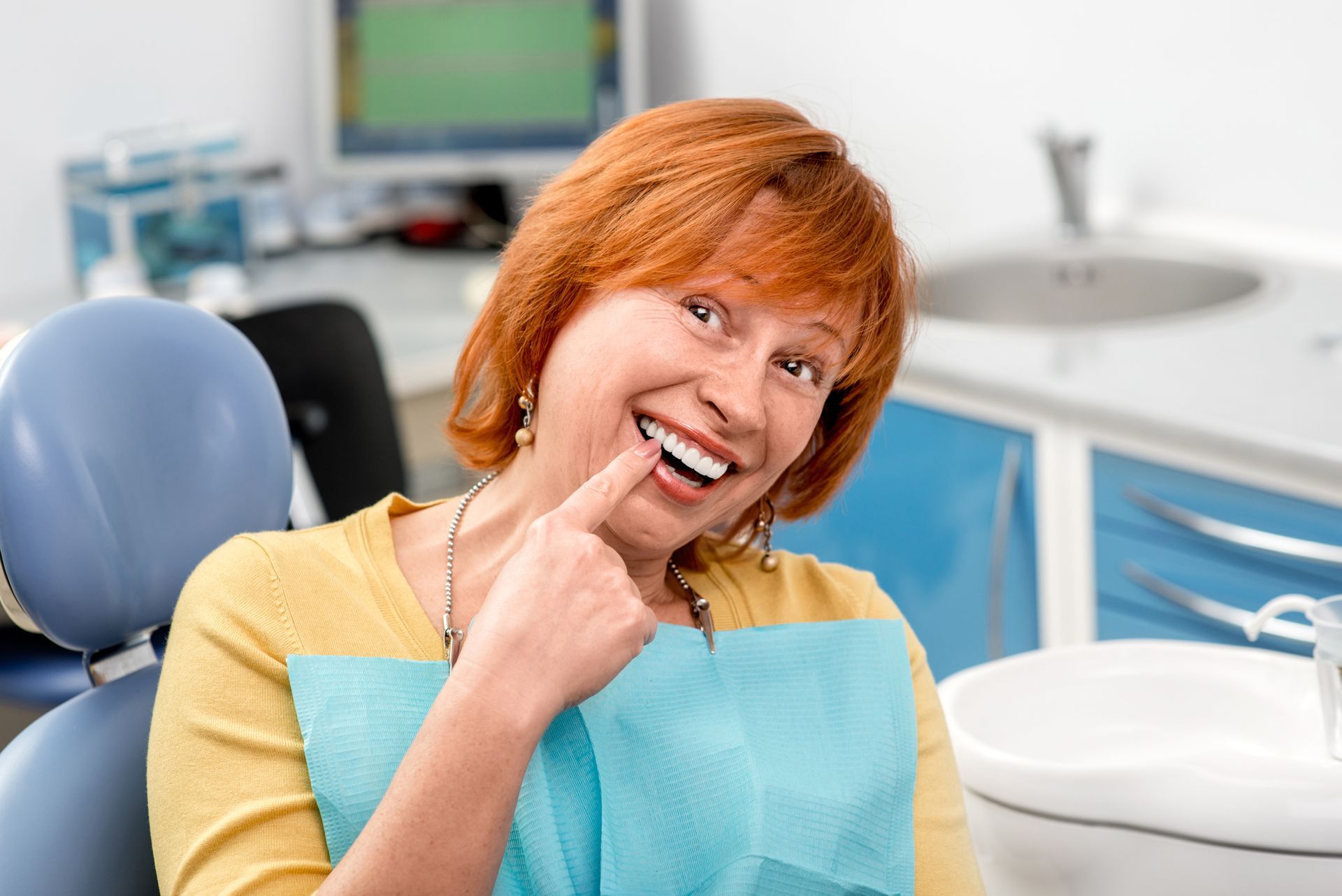 Smiling dental patient in a yellow shirt and blue bib, holding a finger to her cheek in a dentist’s office