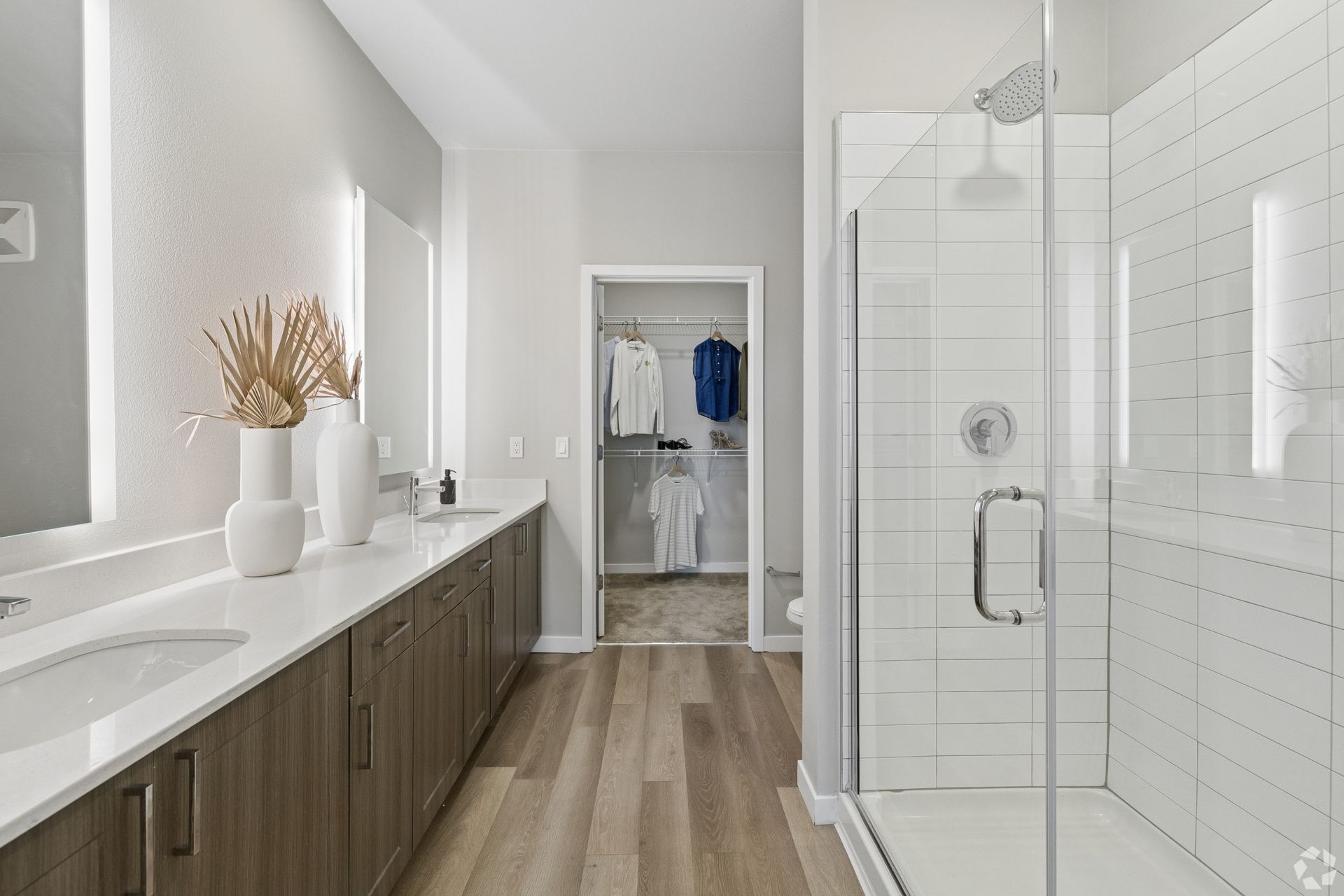 Bright modern bathroom with wood vanity, white countertop, glass shower, and walk-in closet in the background