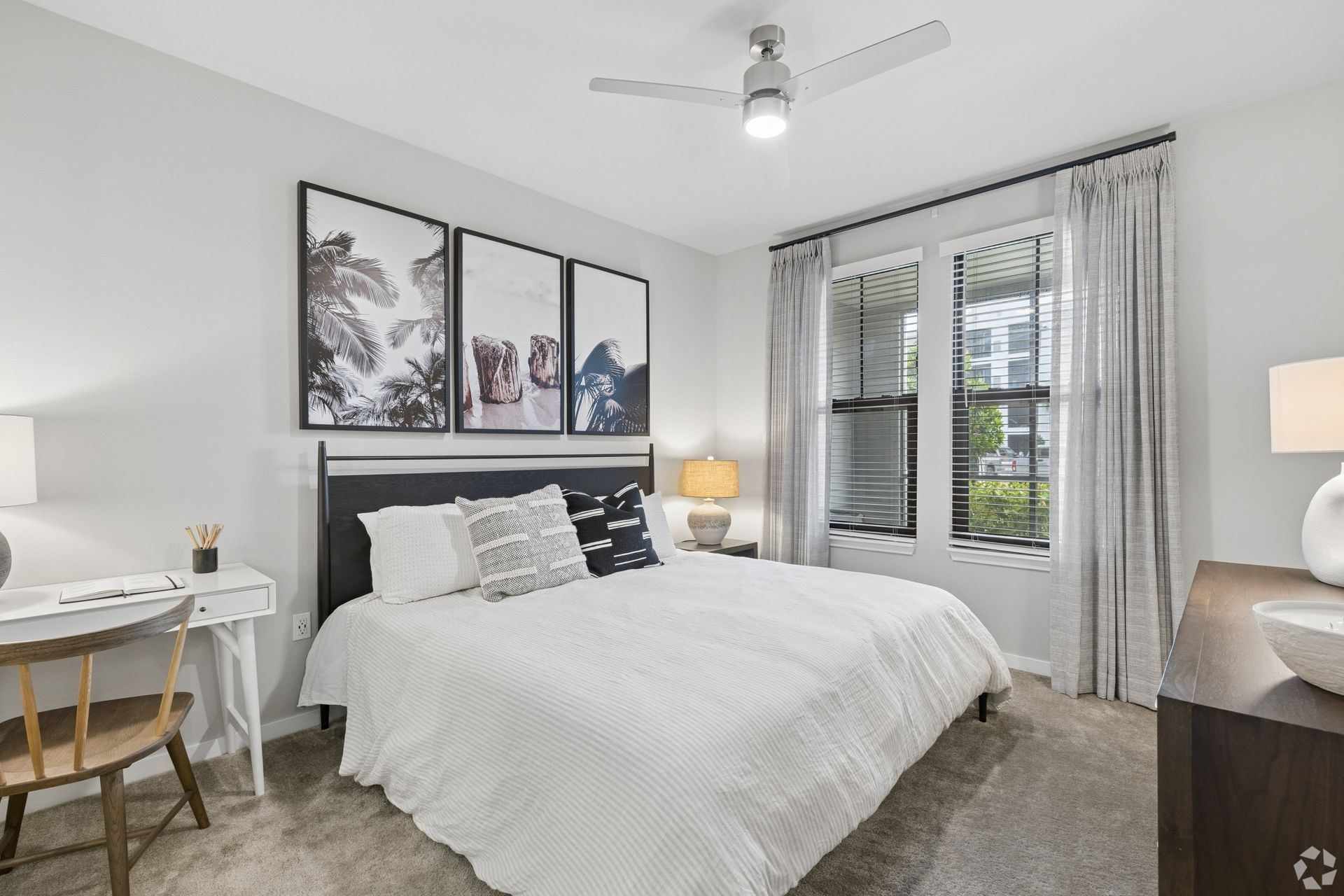 Bright bedroom with a white bed, gray curtains, framed art, and a window with blinds.