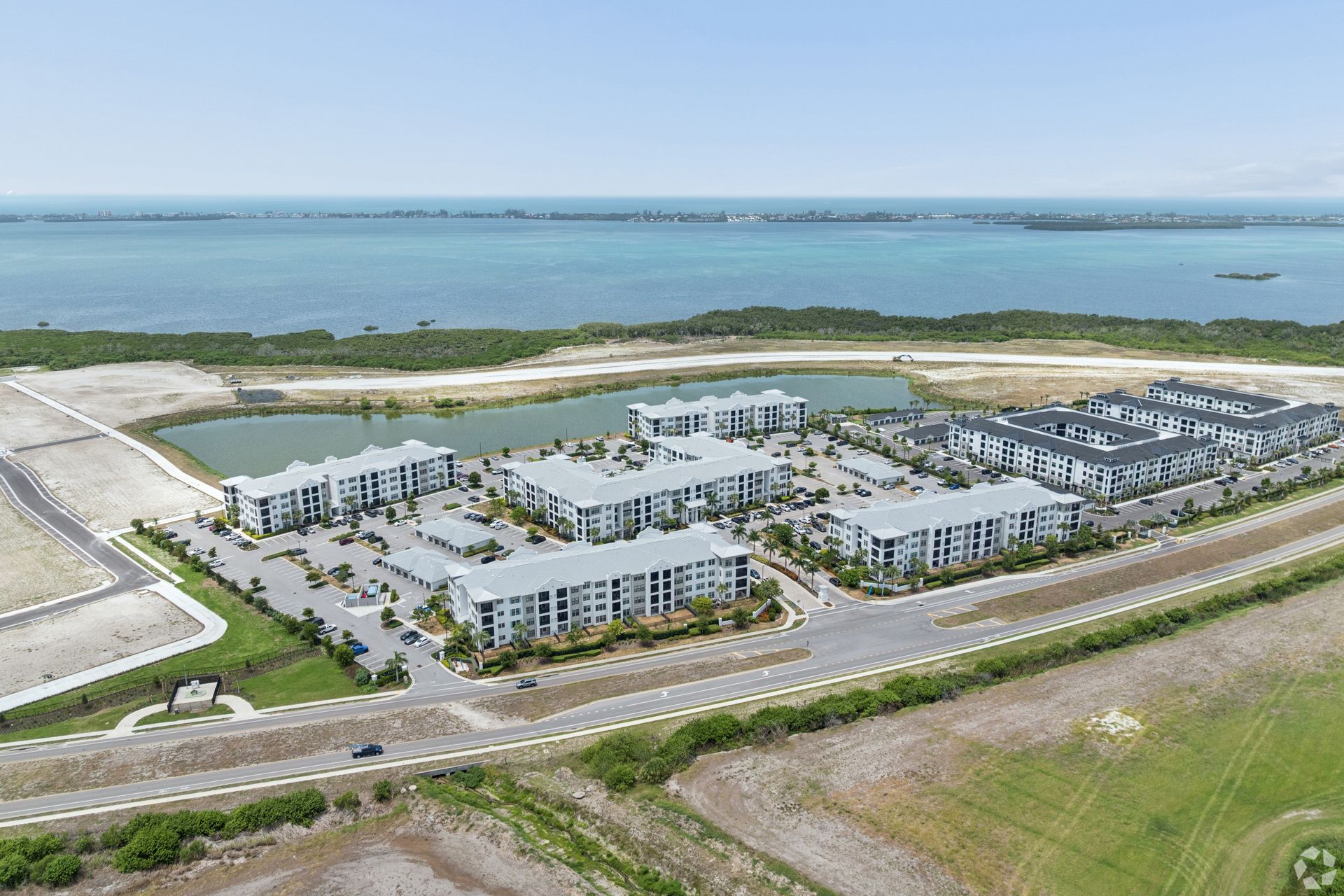 Aerial view of a modern waterfront apartment complex beside a road and blue bay.