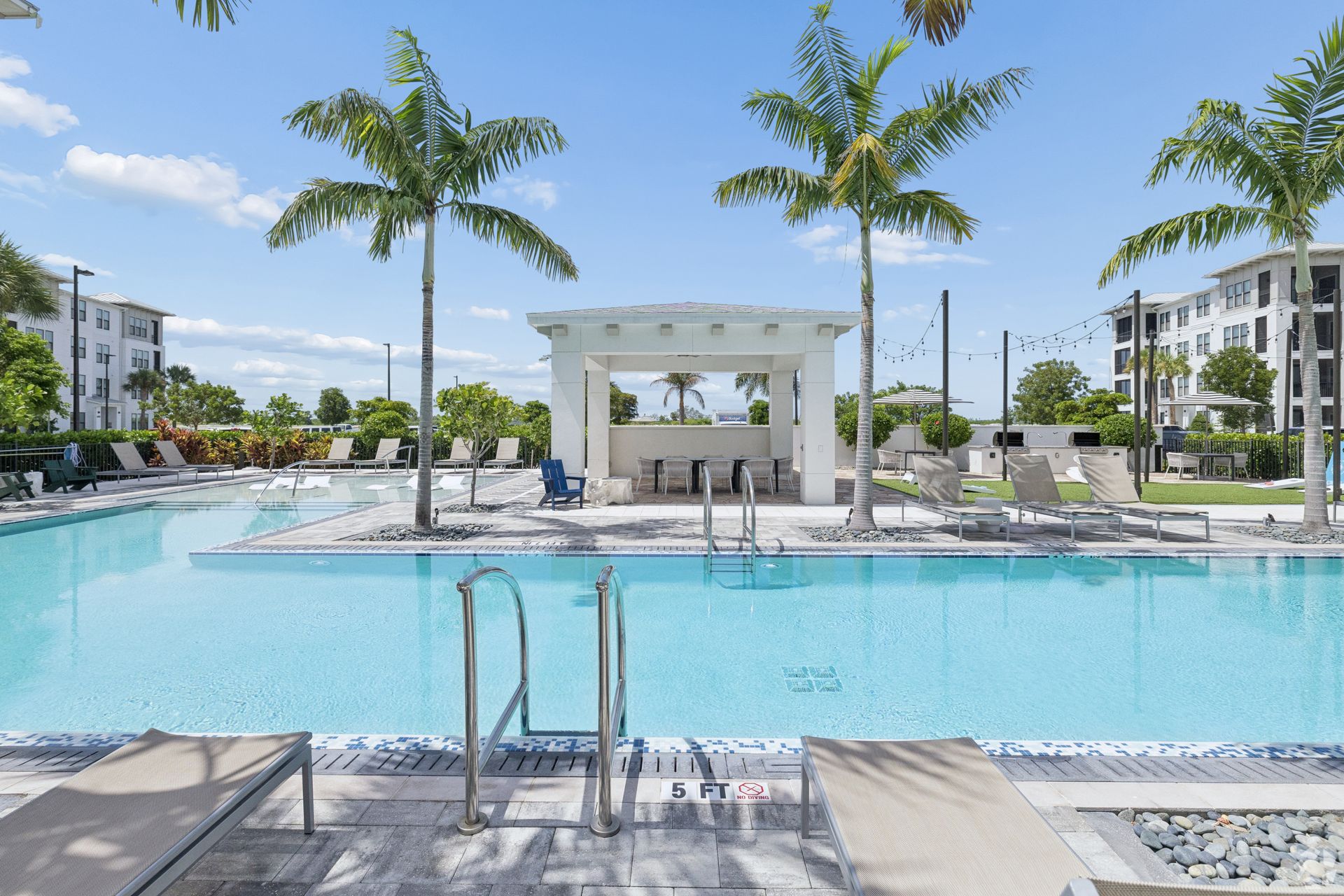 Resort-style pool with lounge chairs, palm trees, and a shaded cabana under a bright blue sky