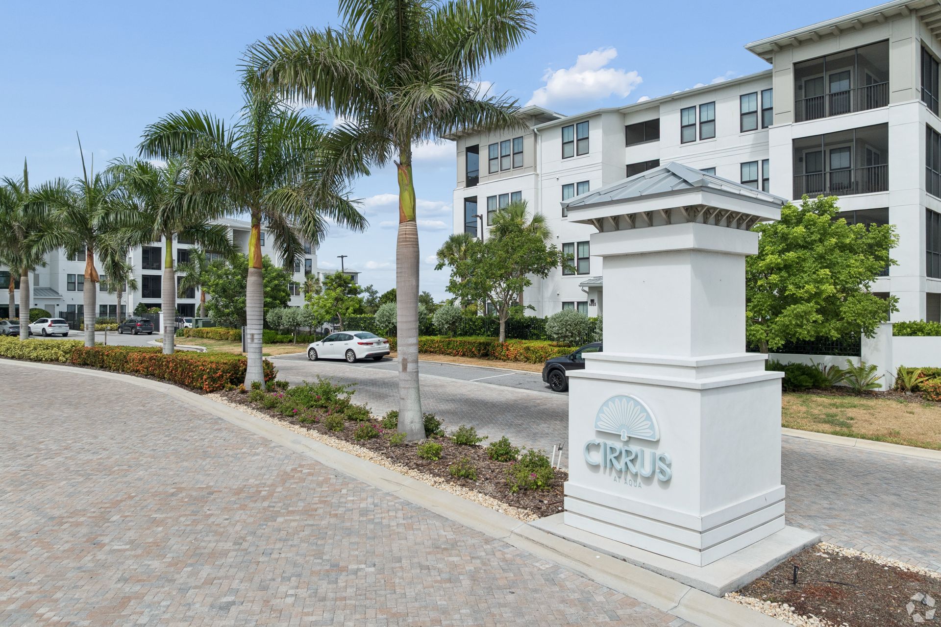 Apartment complex entrance with white sign, palm trees, and parked cars under a blue sky