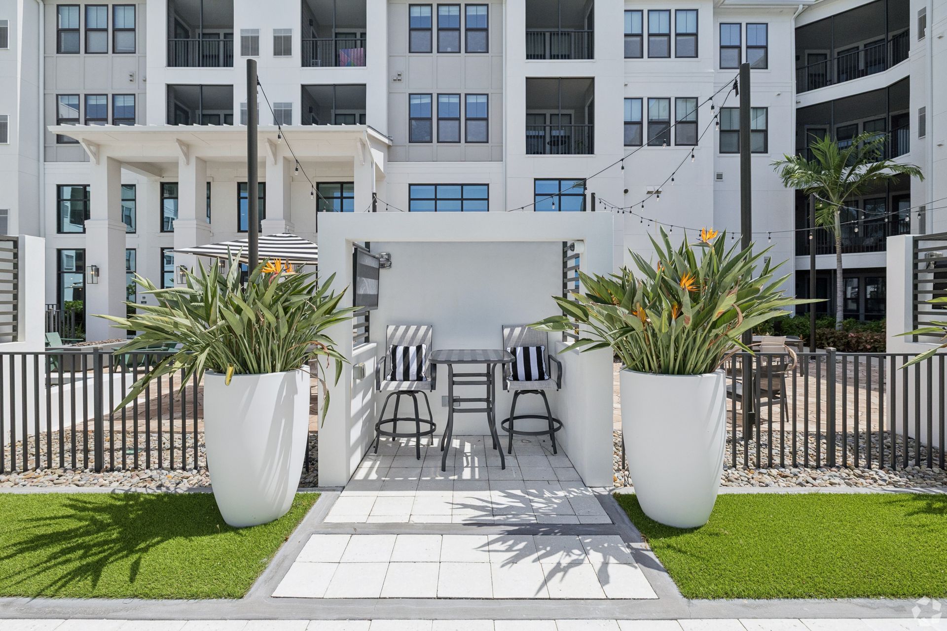 Outdoor patio with white pergola, black chairs, and large potted plants in front of an apartment building