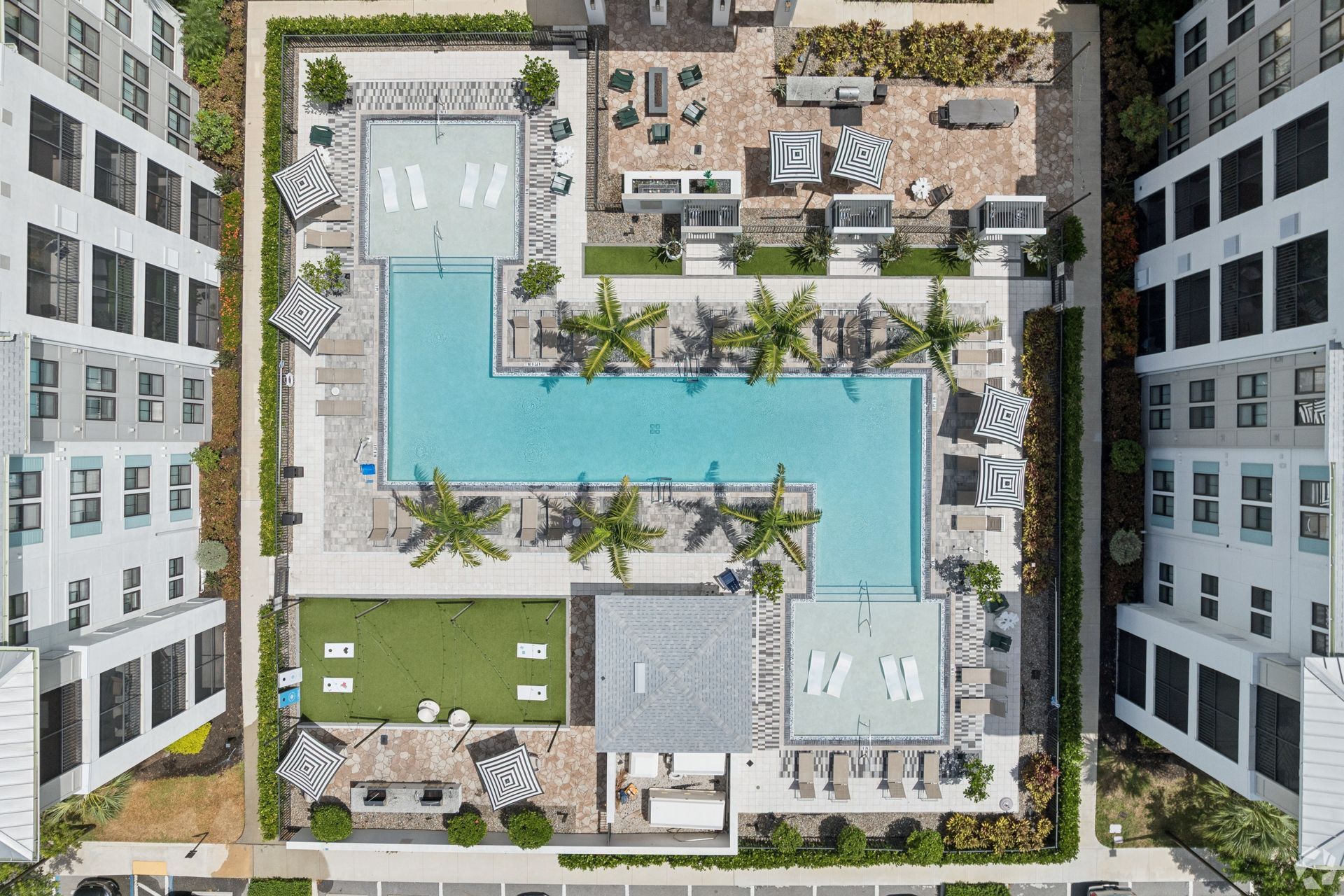 Aerial view of a modern apartment courtyard with a blue L-shaped pool, palm trees, and lounge areas.