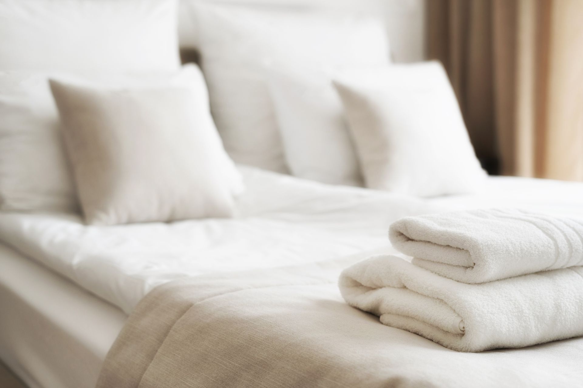 Sunlit bed beside sheer curtains, with a crumpled white blanket and colorful drapes by the window