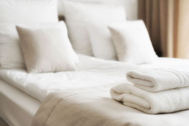 Sunlit bed beside sheer curtains, with a crumpled white blanket and colorful drapes by the window