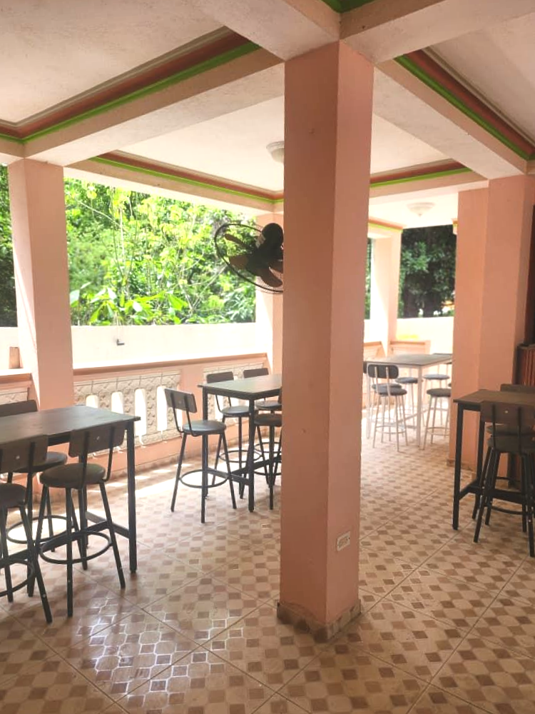 Open-air café with tables and stools on a tiled patio, pink columns, and greenery outside.