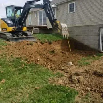 Excavator digging near a house; dirt and grass surround the machine.