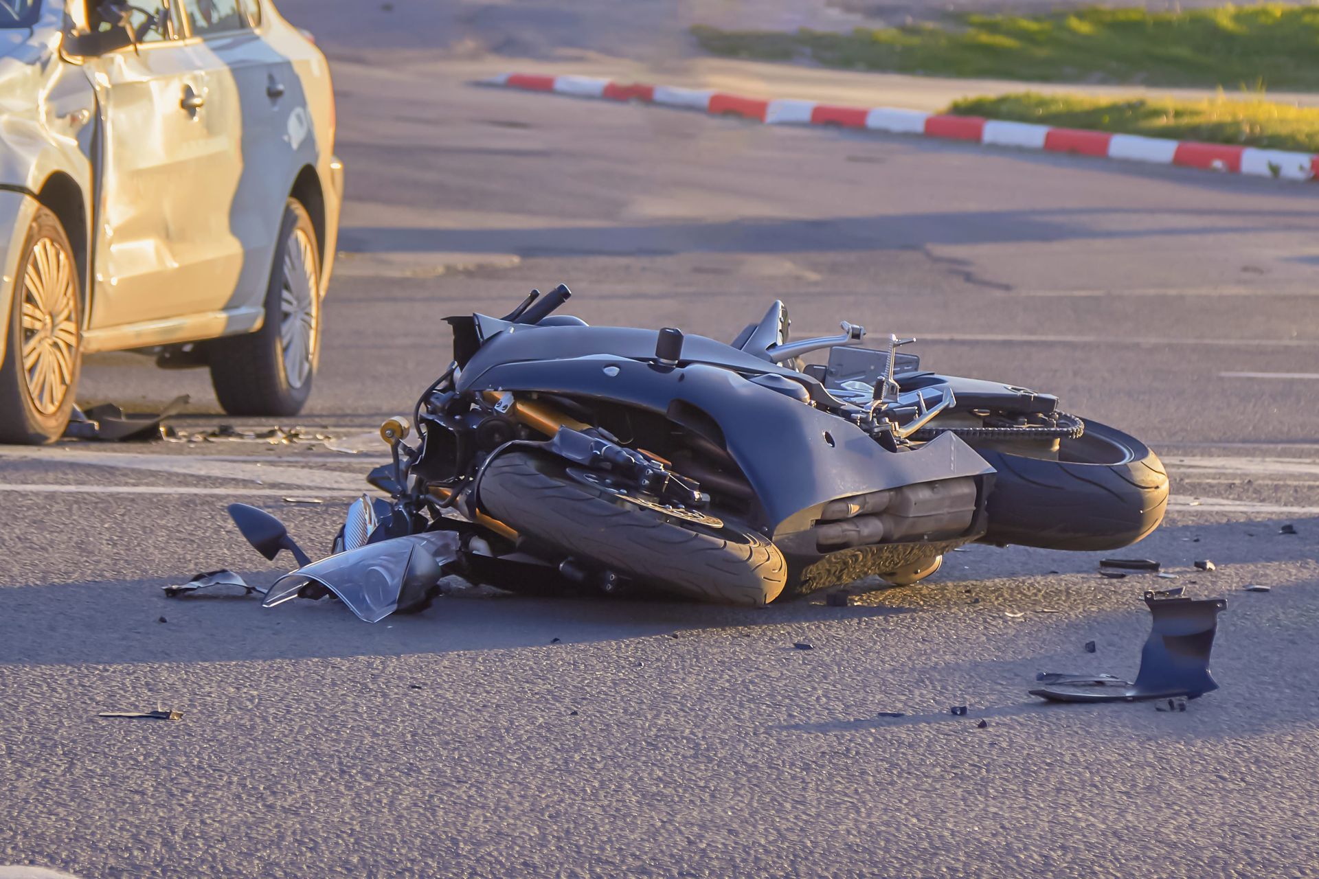 A black motorcycle lies on its side in the middle of a road after an accident, near the front of a damaged silver car.