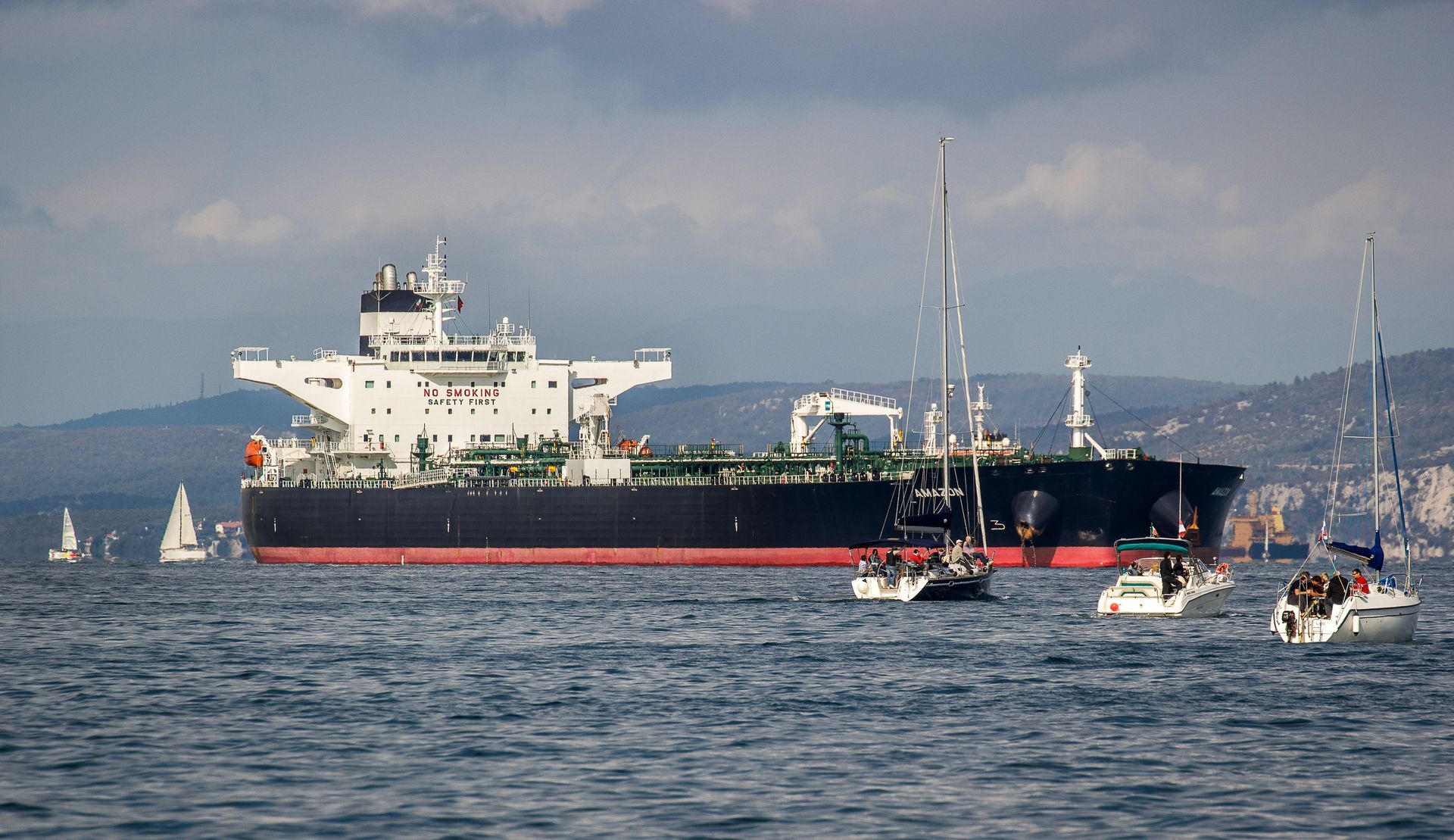 A large black and white cargo ship sailing on the water with several sailboats nearby, set against a hilly coastline.