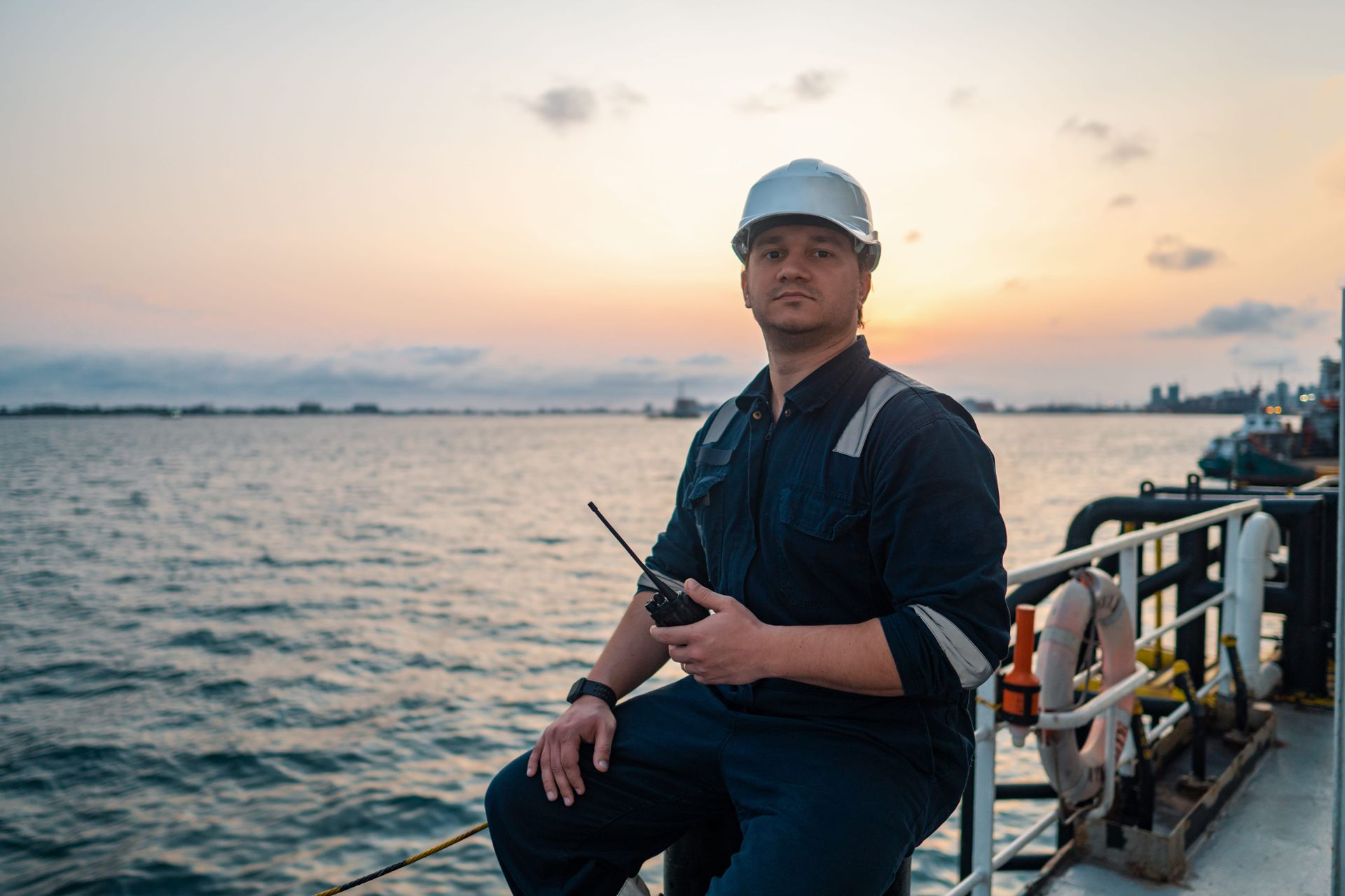 Man standing on a waterfront pier at sunset, holding a radio and wearing a hard hat and dark uniform.