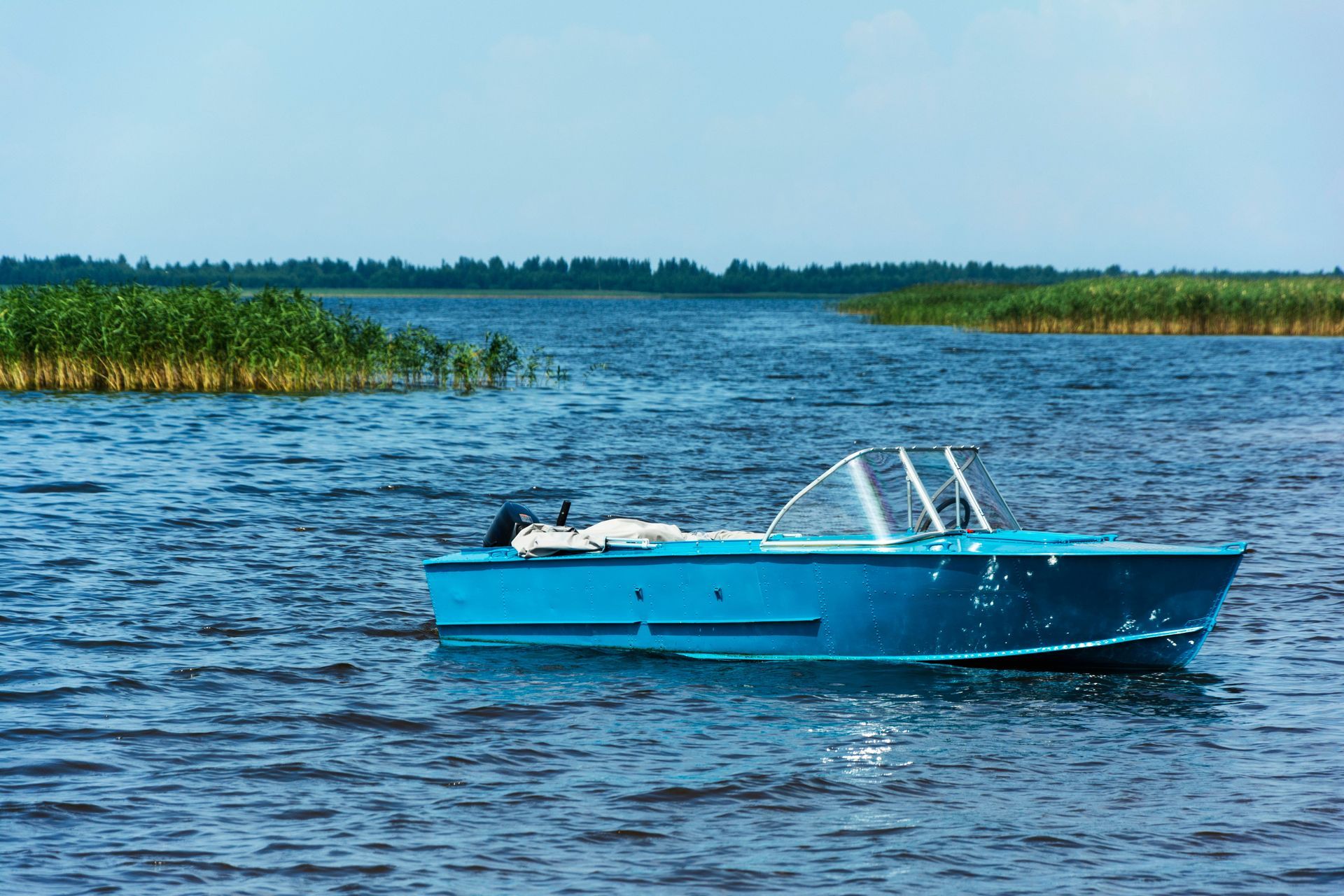 A small, bright blue motorboat floats on a choppy, dark blue lake near grassy shorelines under a clear sky.