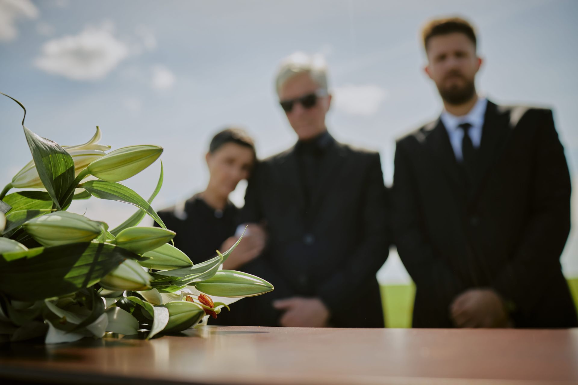 White roses on a coffin with three mourners blurred in the background at an outdoor funeral