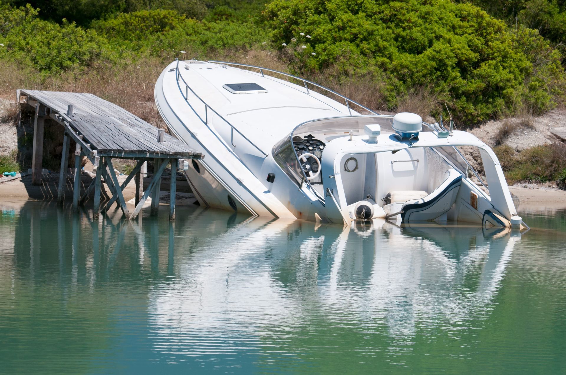 A white motorboat sits partially submerged next to a weathered wooden dock in calm water near a shoreline.