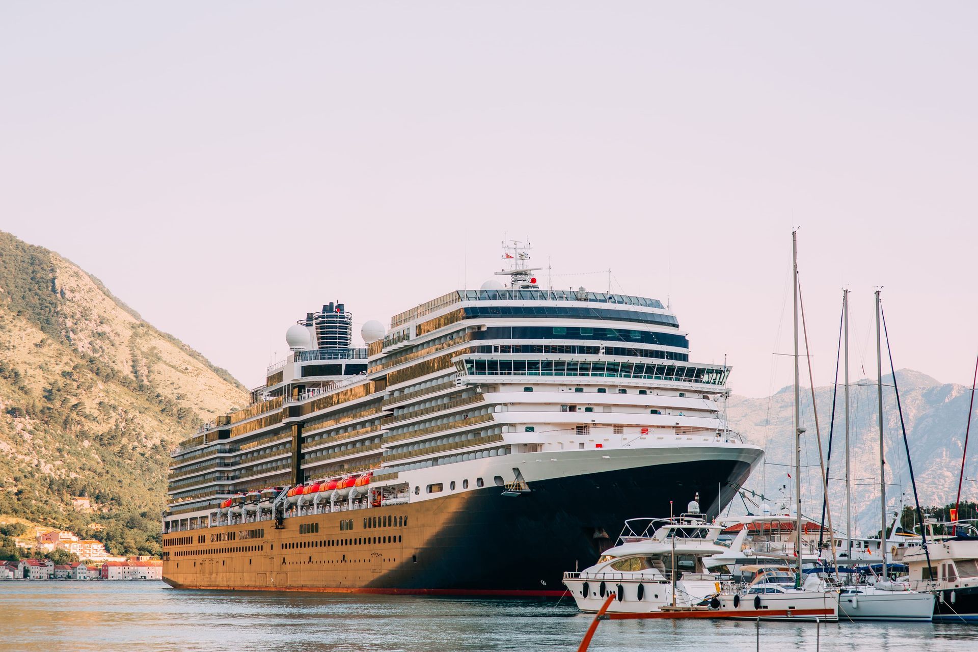 A large cruise ship is docked in a bay near a mountain, with several smaller boats anchored in the foreground.